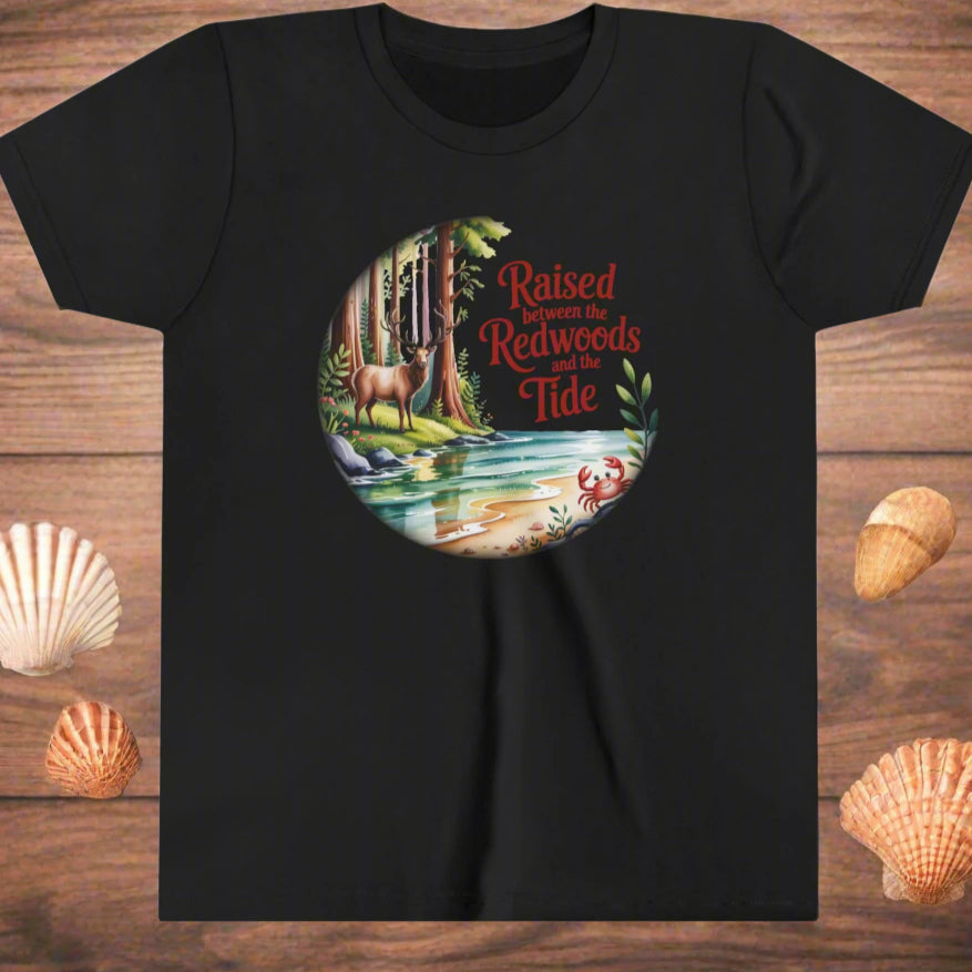 Black t-shirt with nature-themed graphic of redwoods and stream with elk and crab and text Raised between the Redwoods and the Tide.