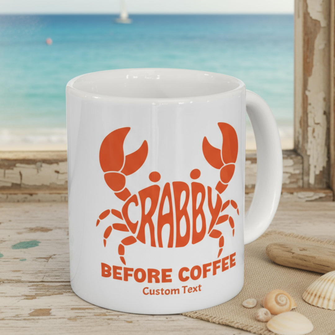 White mug with red crab design and text 'CRABBY BEFORE COFFEE' on a wooden surface with pinecones and a Christmas tree in the background.