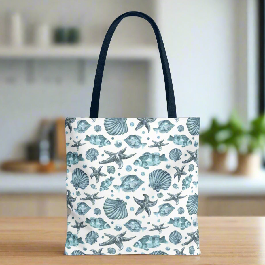 Tote bag with blue strap and an all-over blue seashell, starfish, and fish pattern on a white background with white handles.