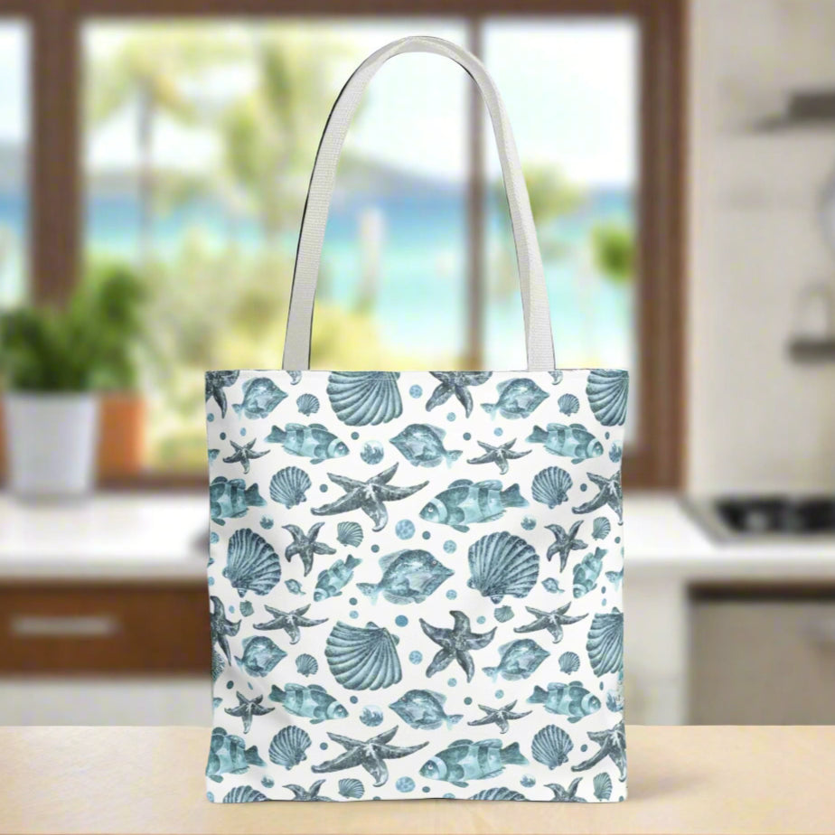 Tote bag with white strap and an all-over blue seashell, starfish, and fish pattern on a white background with white handles.
