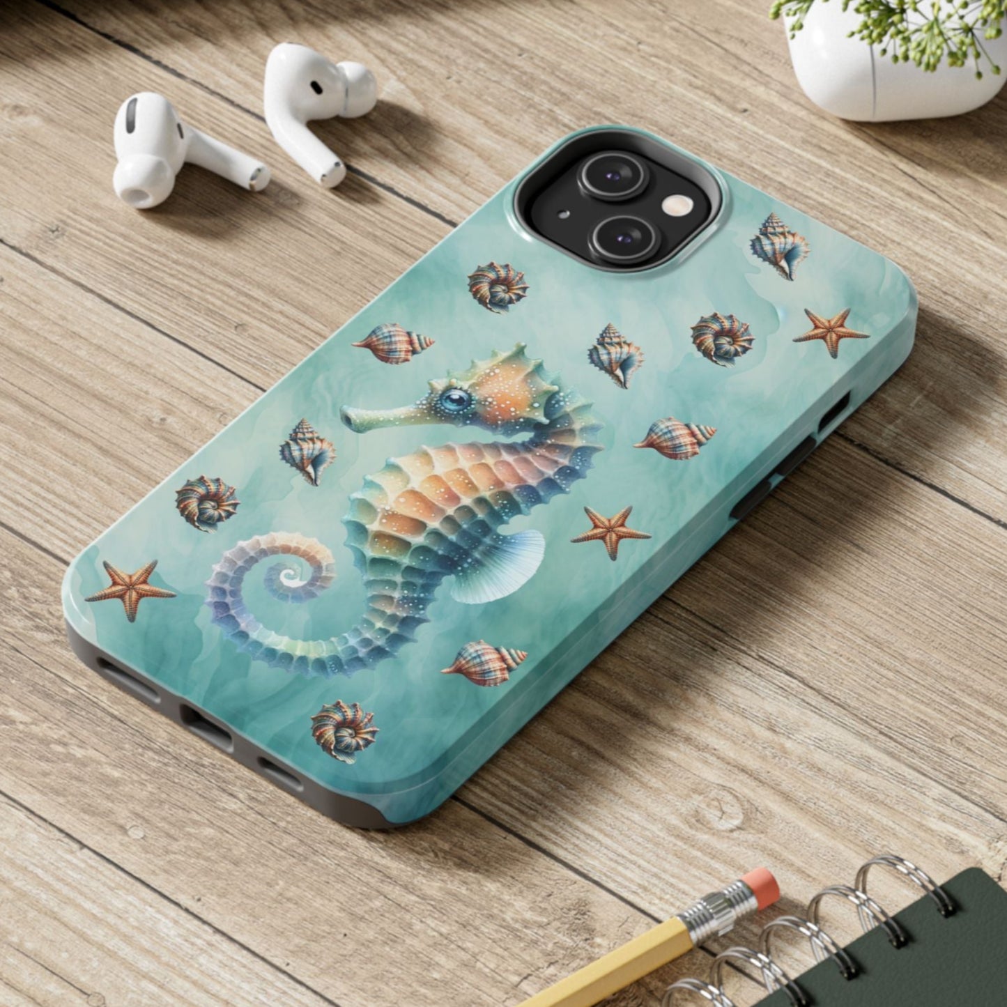 Phone case with seahorse design on a wooden surface with laptop, earbuds, and notebook.