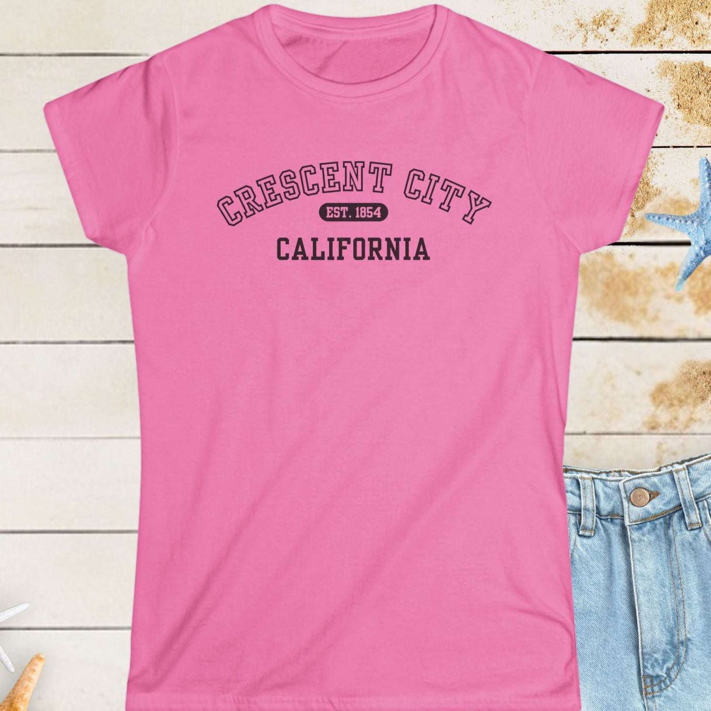 Pink t-shirt with 'Crescent City, California' text on a wooden surface with starfish.
