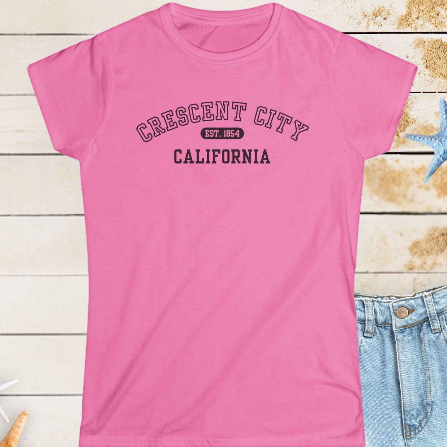 Pink t-shirt with 'Crescent City, California' text on a wooden surface with starfish.