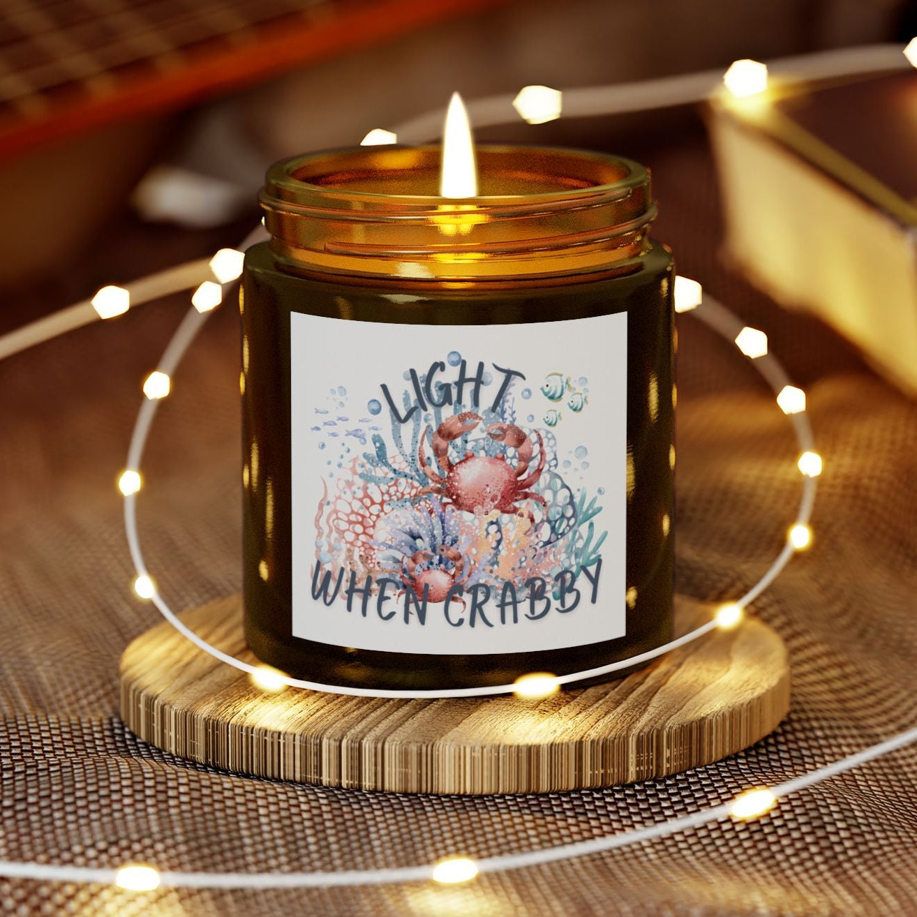 Candle in a jar with a decorative label on a wooden coaster, surrounded by string lights and a guitar in the background.