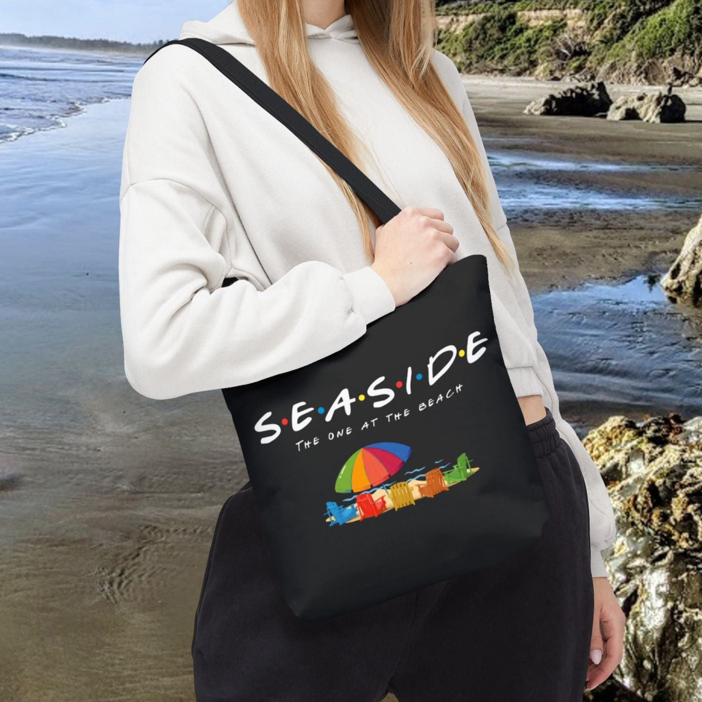 Black 13" x 13" tote bag with the word “Seaside” in rainbow-style letters with colored dots between each letter, underneath says The One at the Beach with five rainbow Adirondack chairs, and a beach umbrella on sand graphic.
