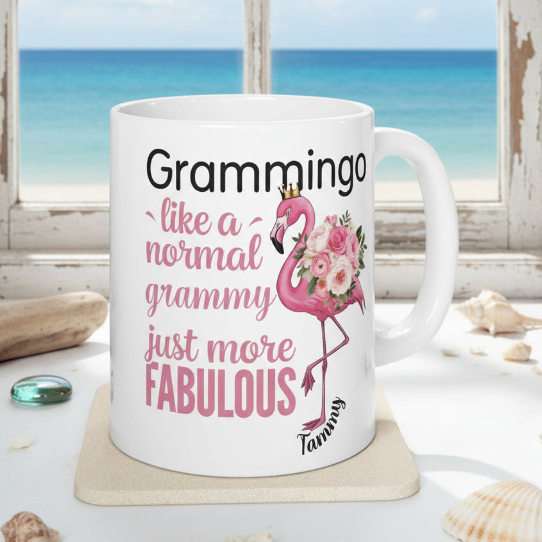 Ceramic coffee mug featuring a flamingo adorned with flowers and the text “Grammingo like a normal Grammy just more fabulous,” available with white, pink, or black handle options.