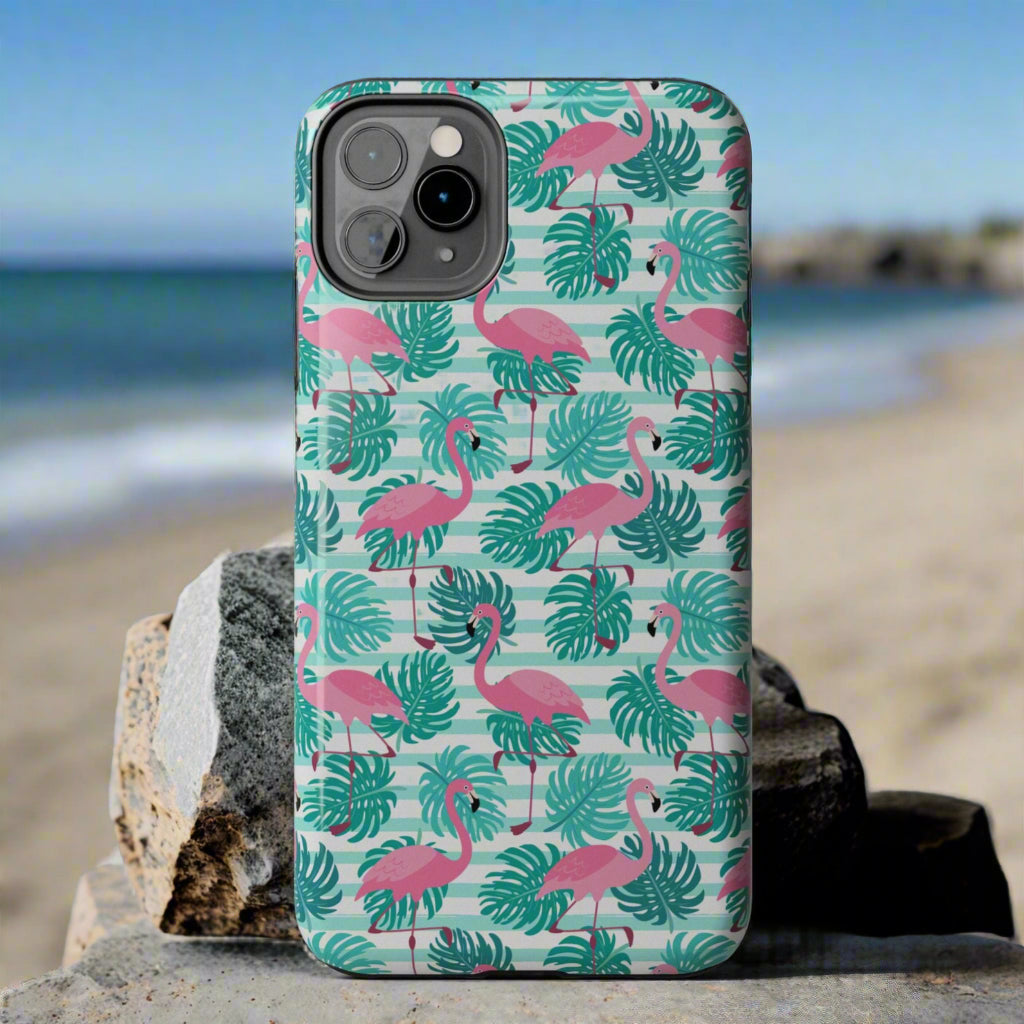 Phone case with pink flamingo and green leaf pattern laying against rocks at the beach.