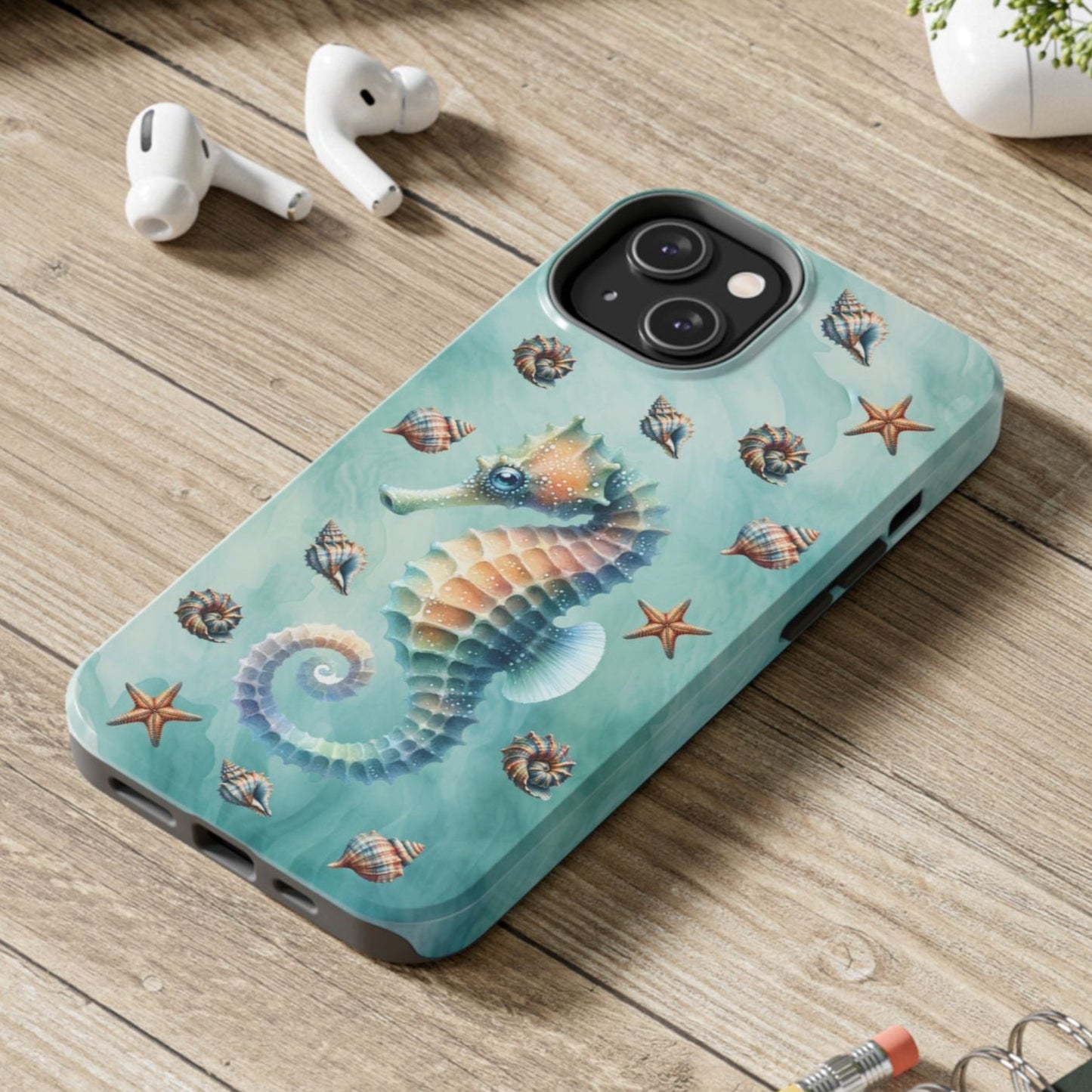 Phone case with seahorse design on a wooden surface with laptop, earbuds, and notebook.