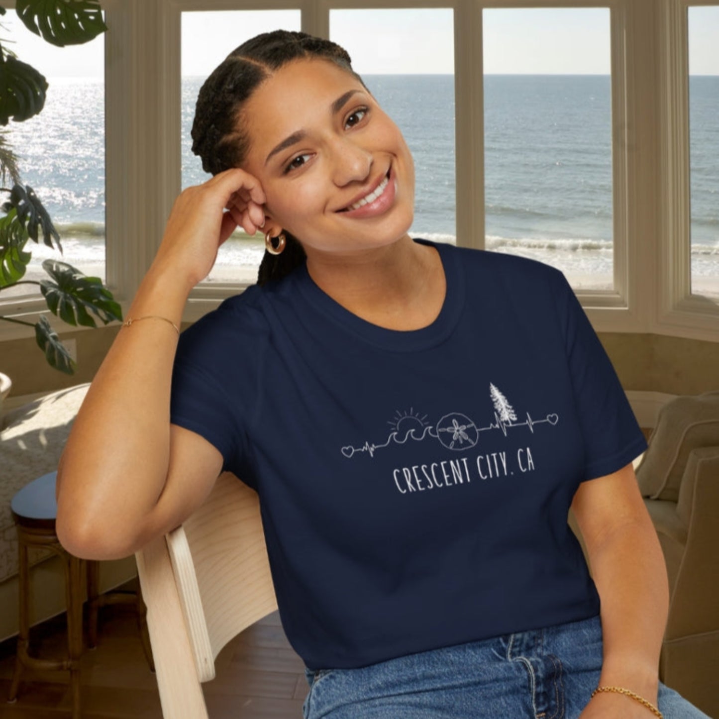 Woman wearing a navy blue t-shirt with 'Crescent City, CA' design sitting in a room with large windows overlooking the ocean.
