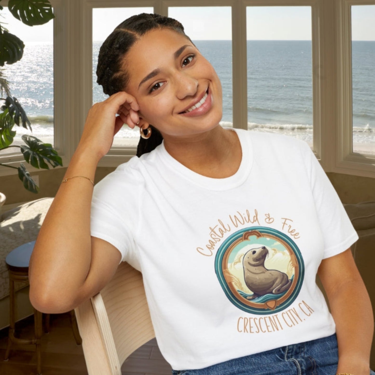 Woman wearing a white t-shirt with a graphic design sitting in a room with large windows showing an ocean view.