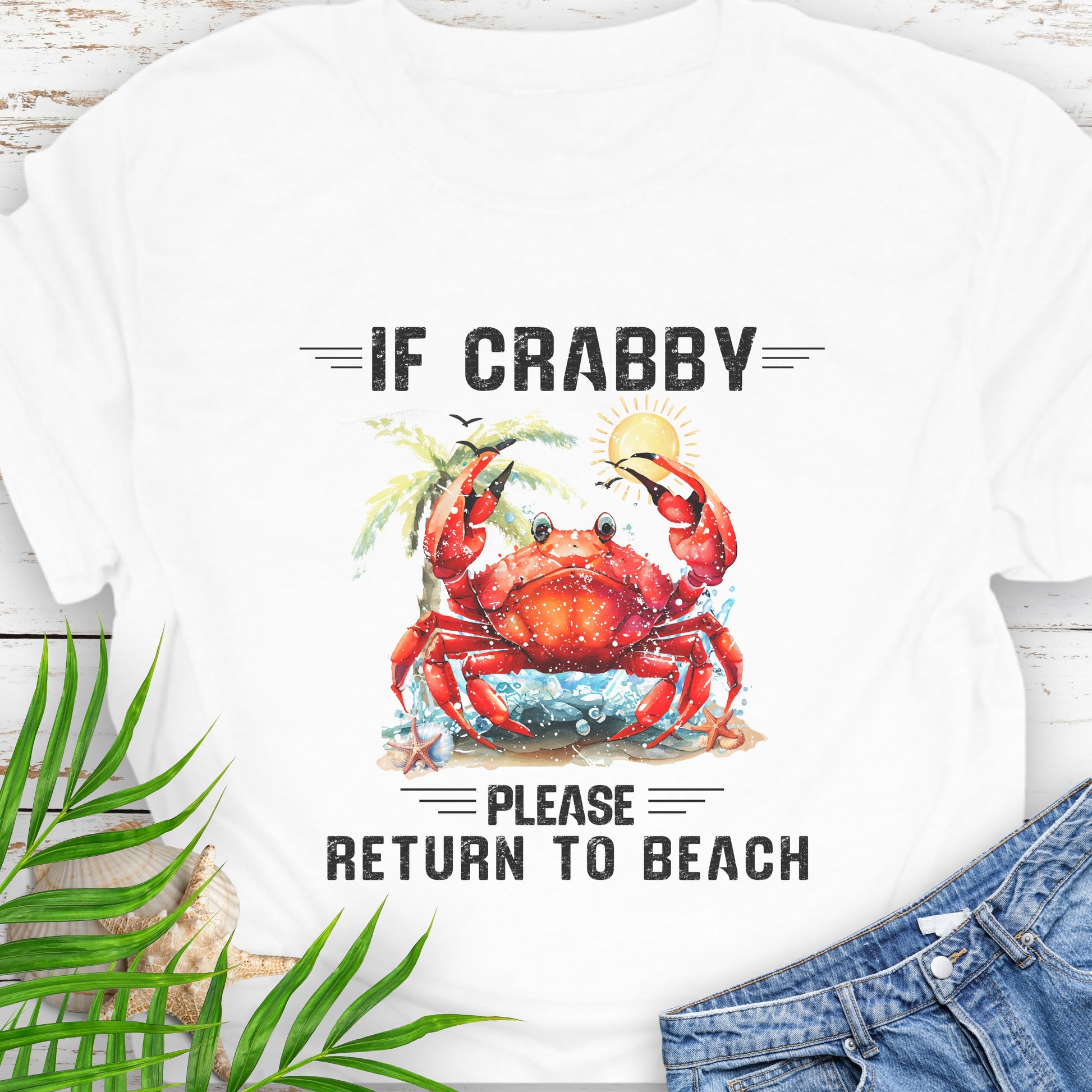 White t-shirt with a red crab graphic and text If Crabby Please Return to Beach on a wooden surface with leaves and jeans.