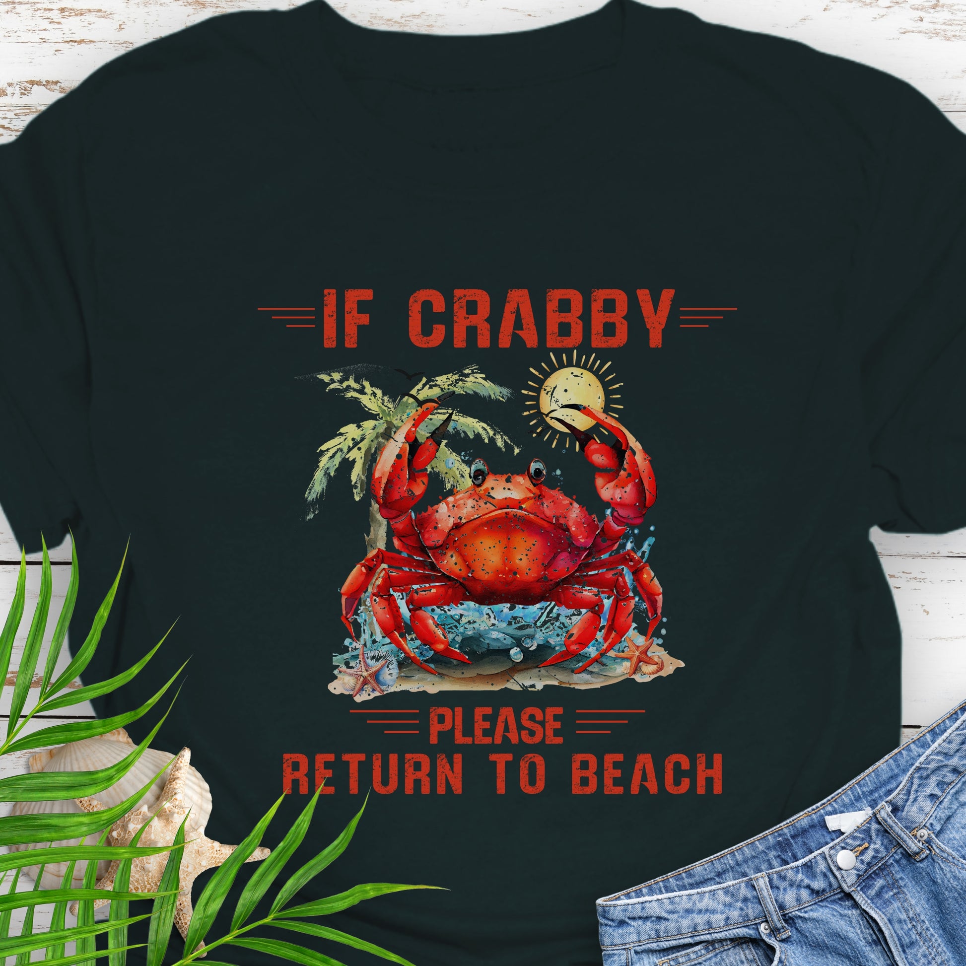 Black t-shirt with a red crab graphic and text If Crabby Please Return to Beach on a wooden surface with leaves and jeans.