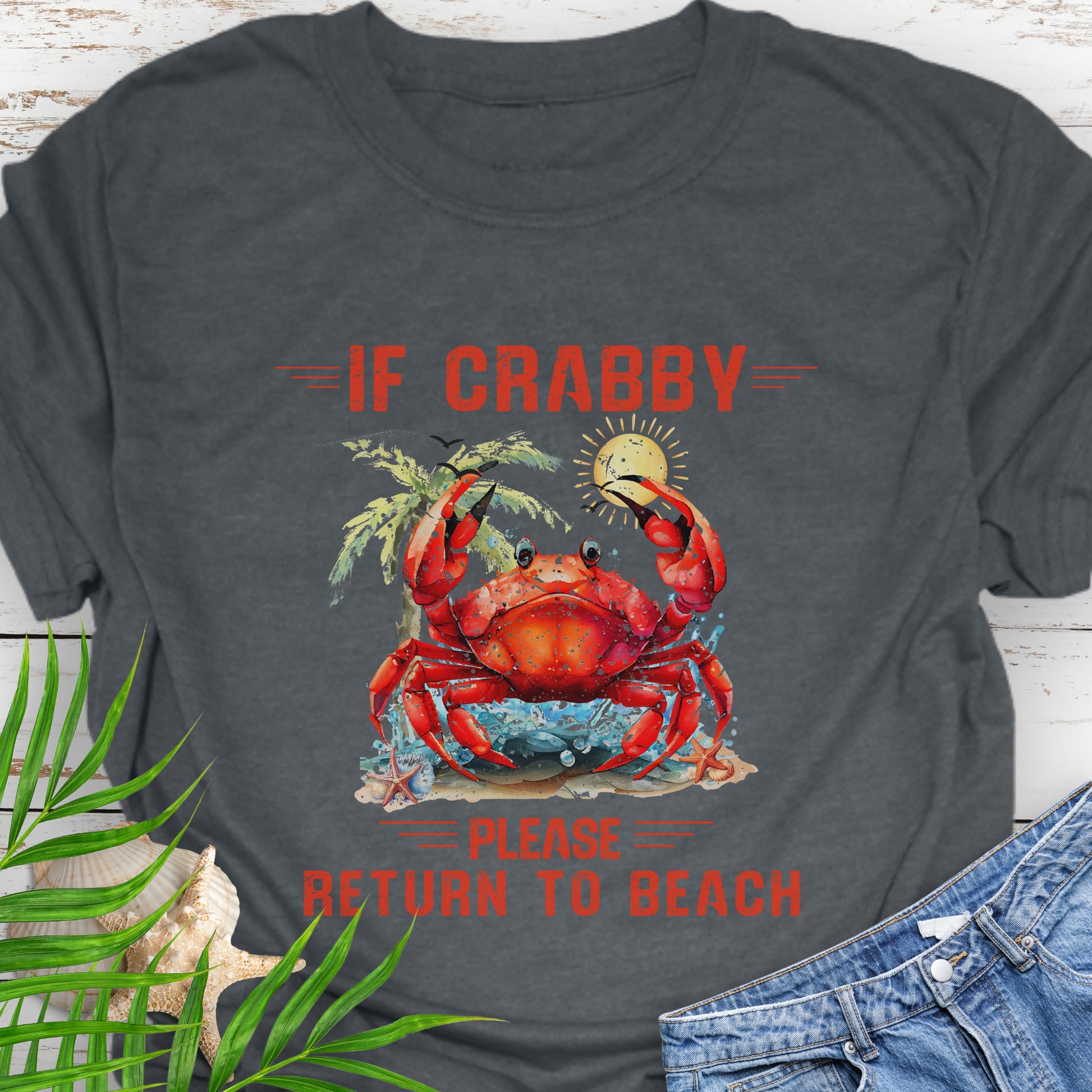 Dark Heather Gray t-shirt with a red crab graphic and text If Crabby Please Return to Beach on a wooden surface with leaves and jeans.