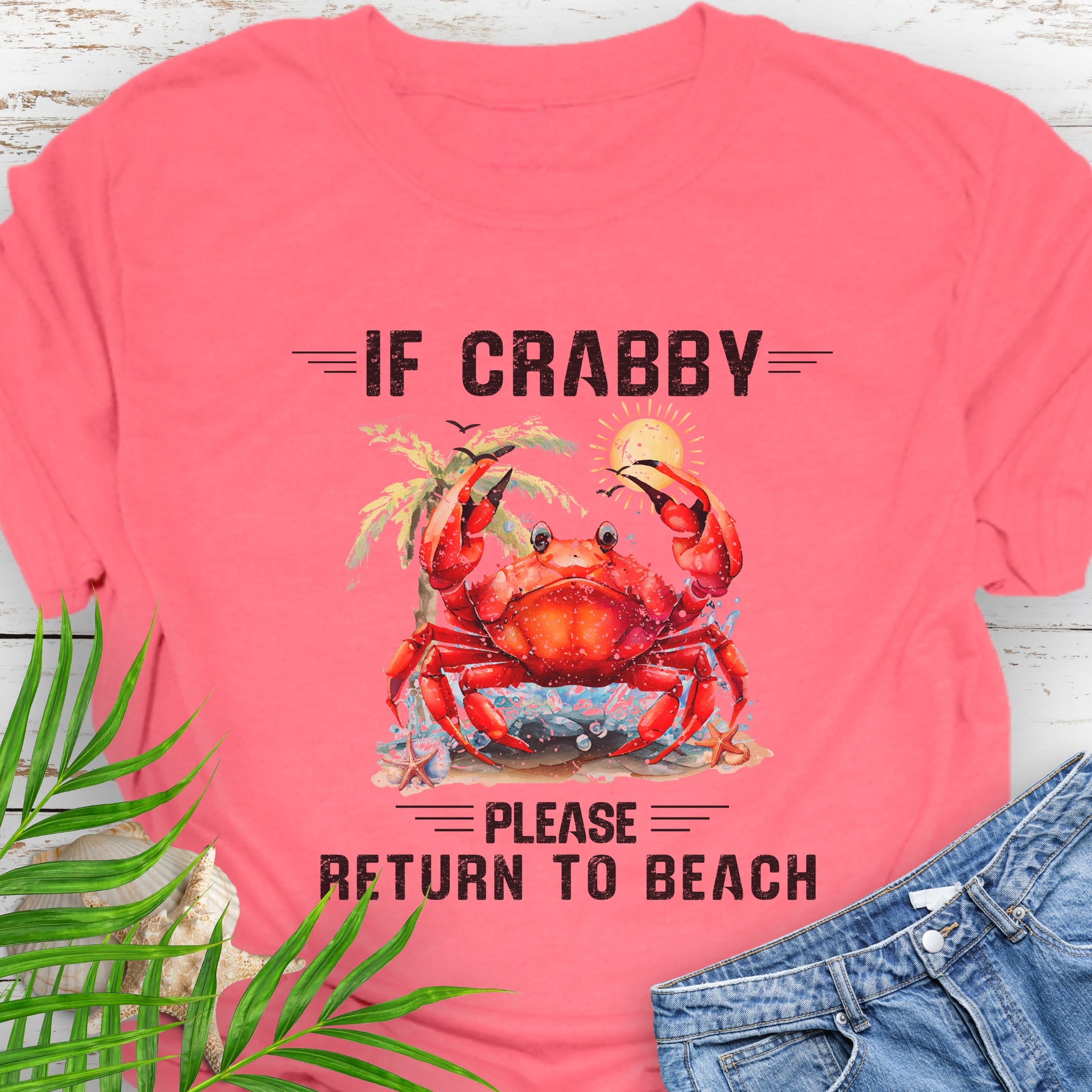 Coral Silk t-shirt with a red crab graphic and text If Crabby Please Return to Beach on a wooden surface with leaves and jeans.