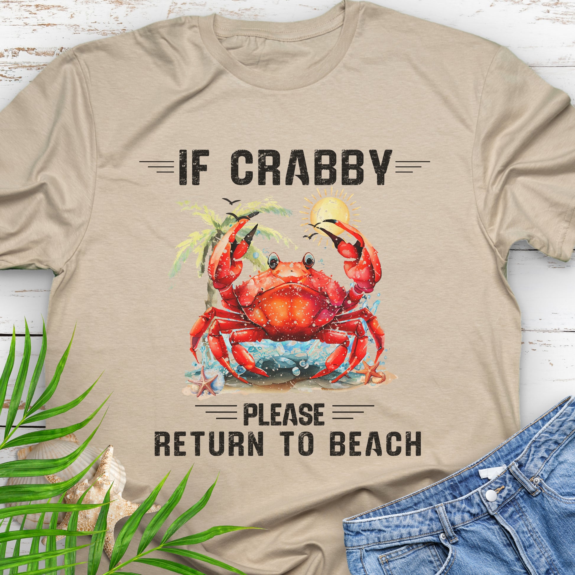 Sand beige t-shirt with a red crab graphic and text If Crabby Please Return to Beach on a wooden surface with leaves and jeans.