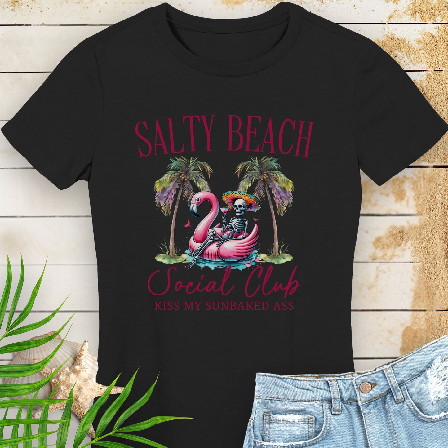 Black women’s t-shirt with skeleton in beach outfit floating in a flamingo pool float with palm trees and text “Salty Beach Social Club – Kiss My Sunbaked Ass.”