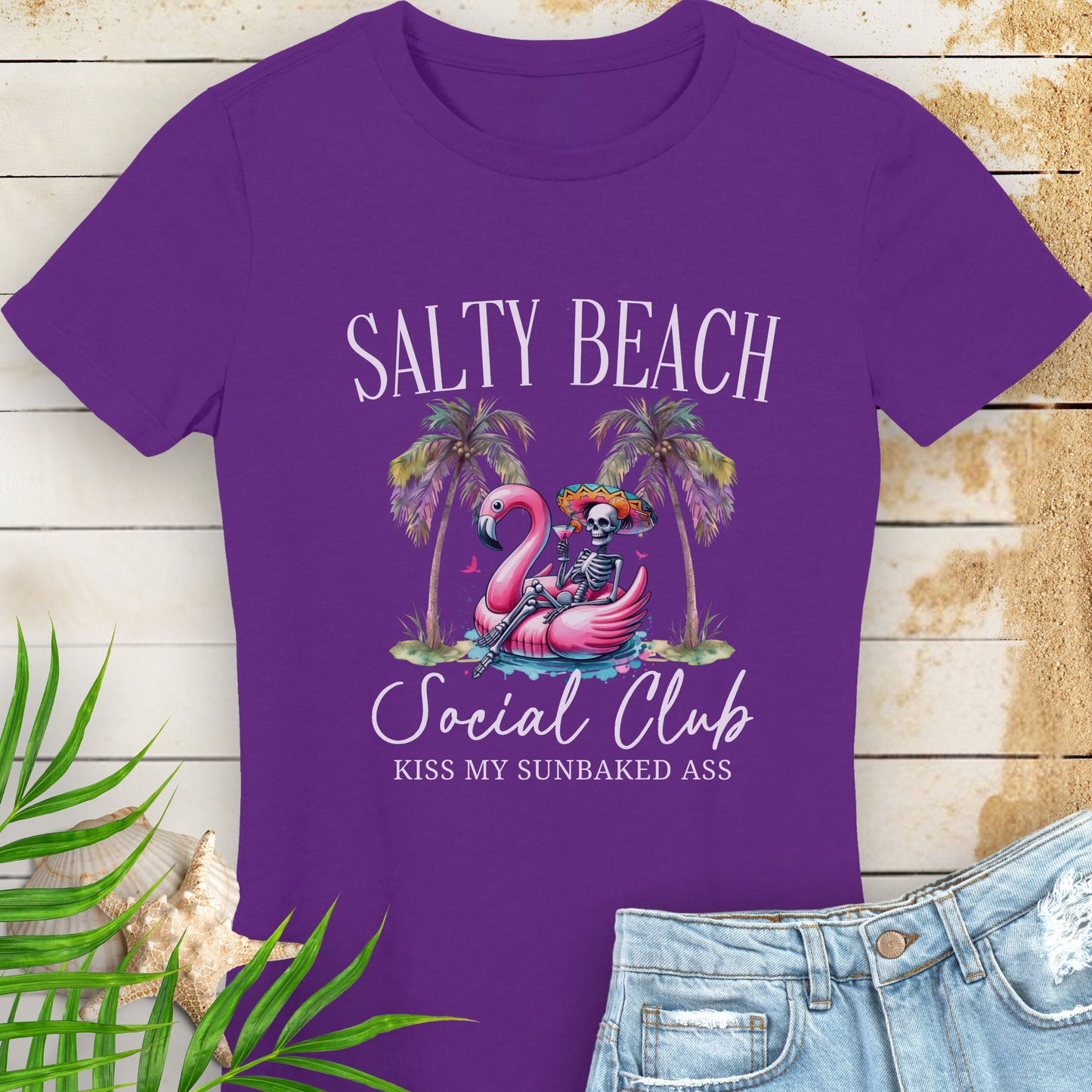 Purple women’s t-shirt with skeleton in beach outfit floating in a flamingo pool float with palm trees and text “Salty Beach Social Club – Kiss My Sunbaked Ass.”