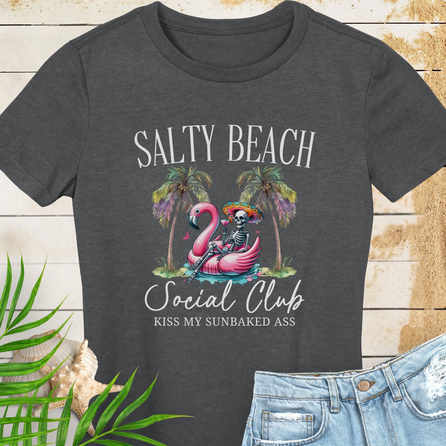 Dark Gray women’s t-shirt with skeleton in beach outfit floating in a flamingo pool float with palm trees and text “Salty Beach Social Club – Kiss My Sunbaked Ass.”