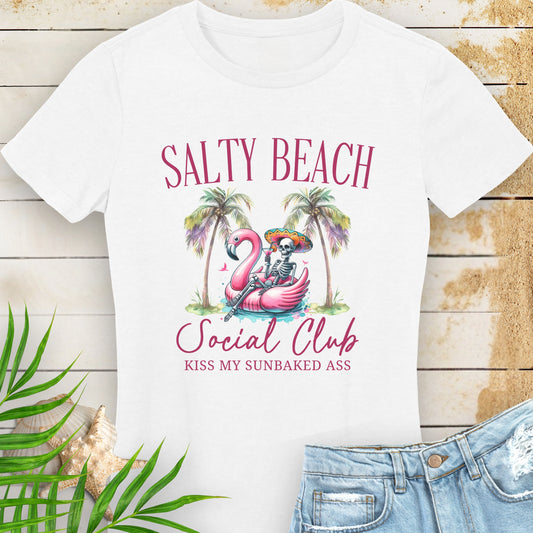 White women’s t-shirt with skeleton in beach outfit floating in a flamingo pool float with palm trees and text “Salty Beach Social Club – Kiss My Sunbaked Ass.”