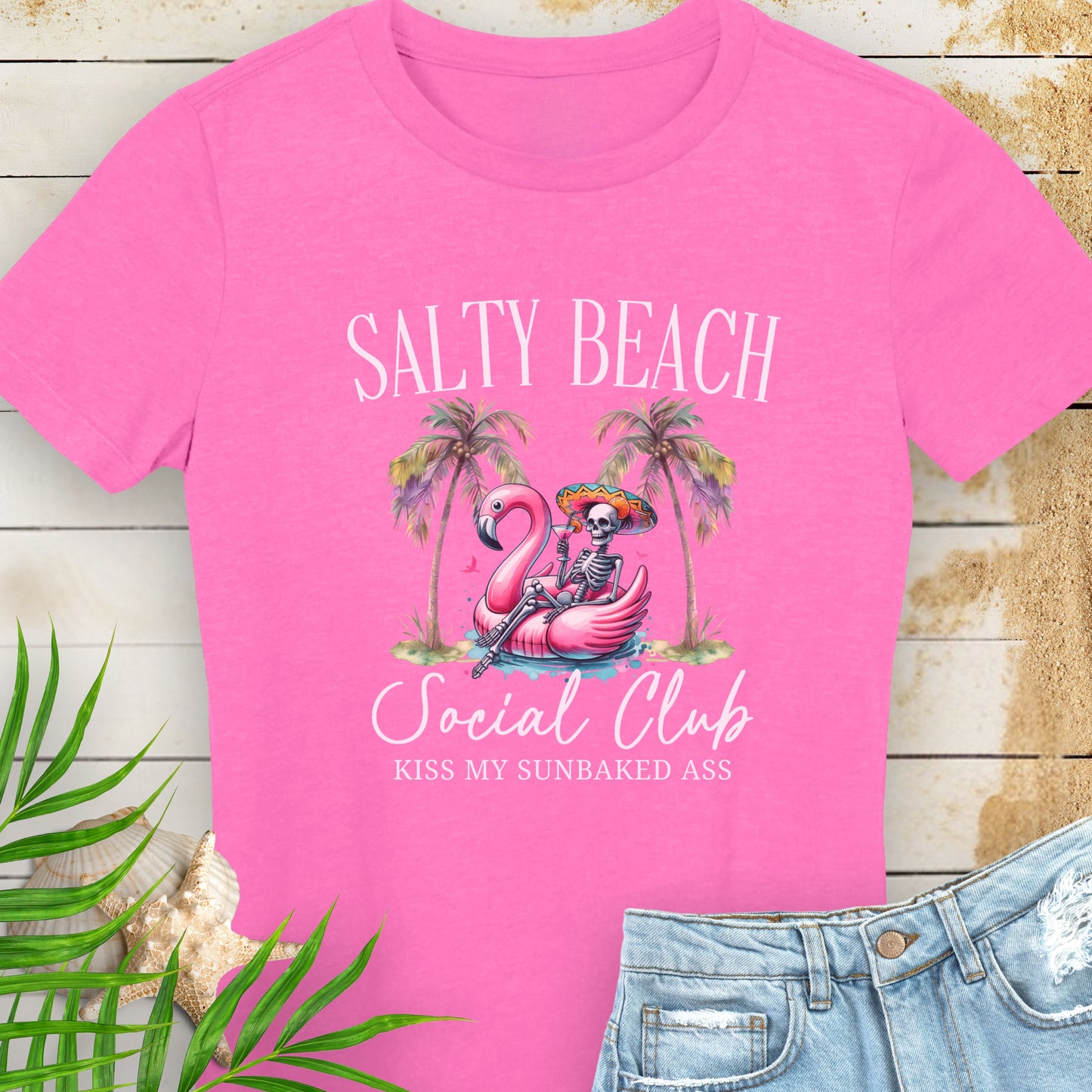 Pink women’s t-shirt with skeleton in beach outfit floating in a flamingo pool float with palm trees and text “Salty Beach Social Club – Kiss My Sunbaked Ass.”