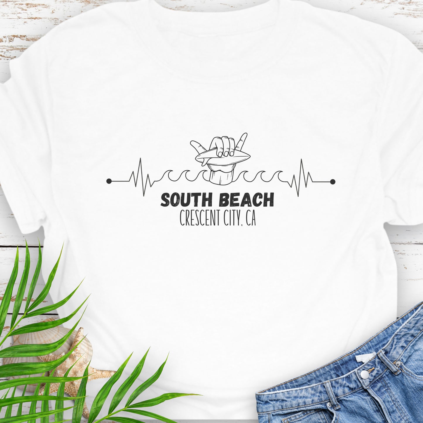 White t-shirt with 'South Beach Crescent City, CA' text with heartline of waves and a hang ten hand holding a surfboard.