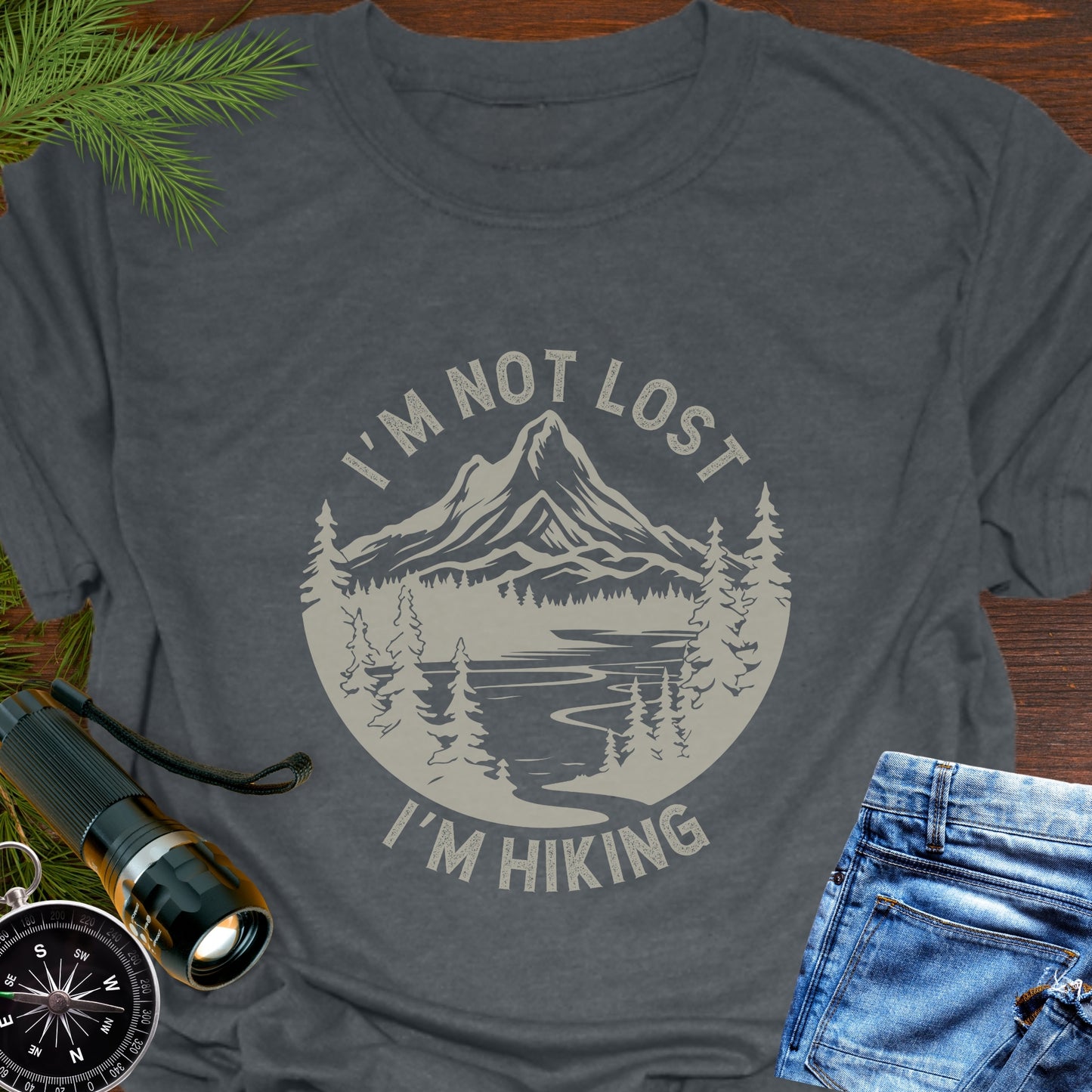 Gray t-shirt with 'I'm not lost, I'm hiking' design on a wooden surface with outdoor accessories.