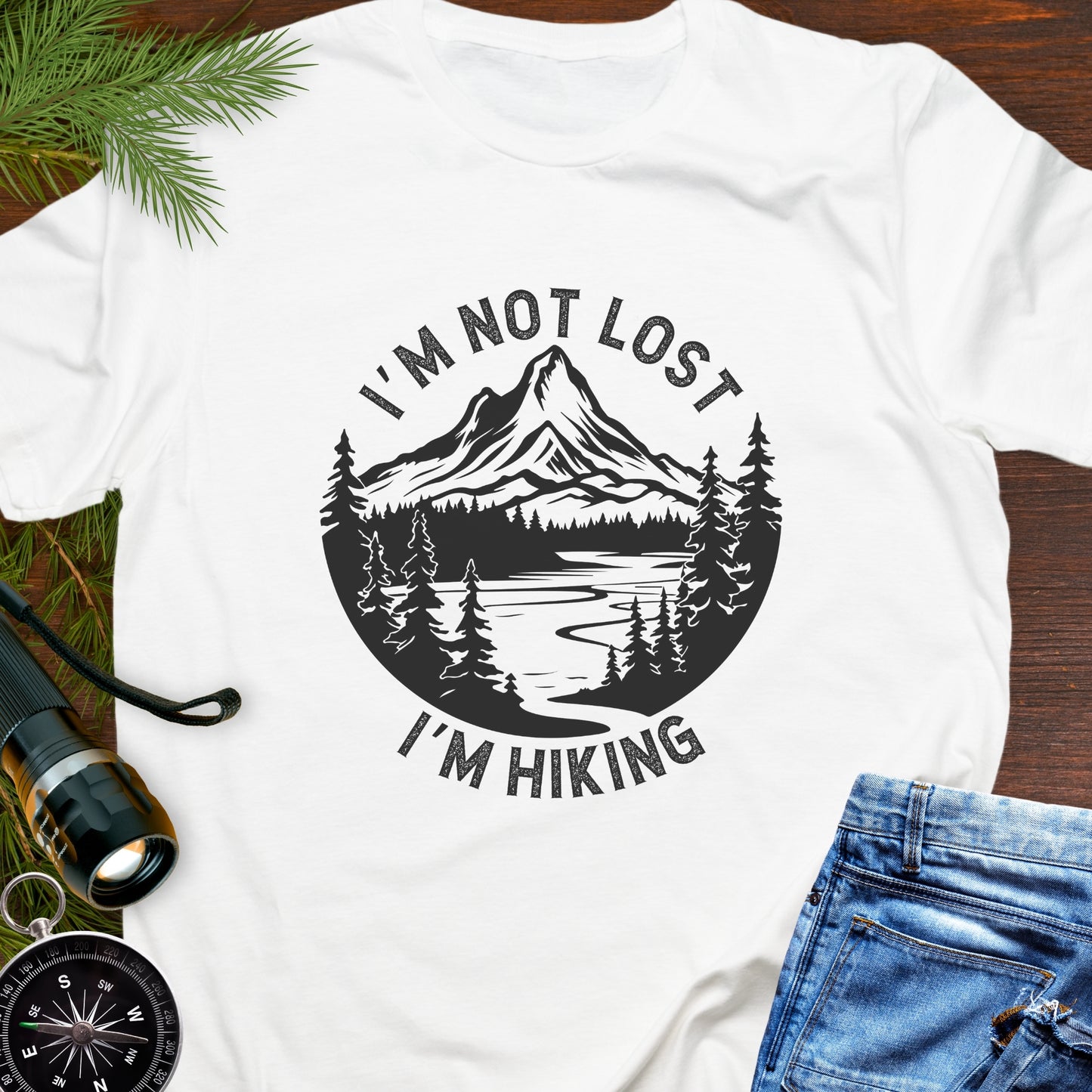 White t-shirt with 'I'm not lost, I'm hiking' design on a wooden surface with nature-themed accessories.