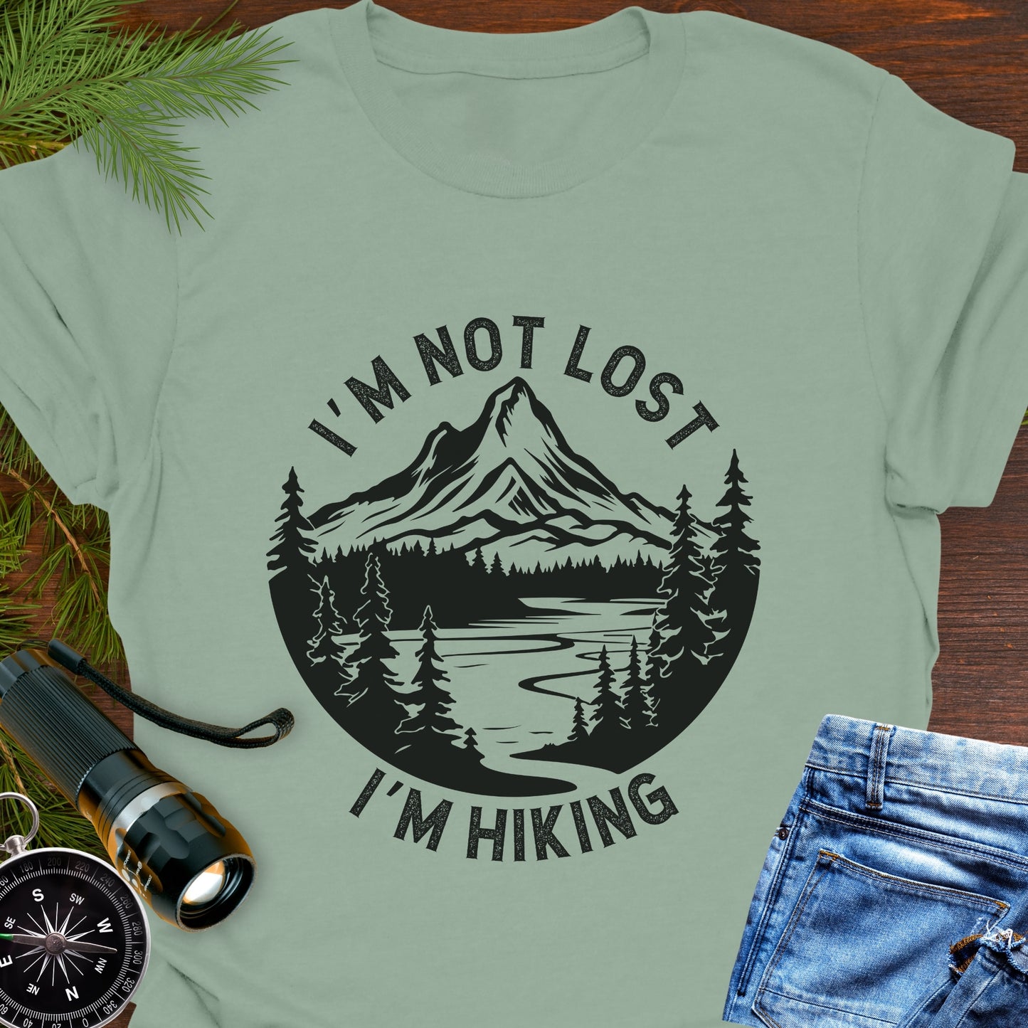 Light green t-shirt with mountain graphic and text 'I'm not lost, I'm hiking' on a wooden surface with outdoor accessories.