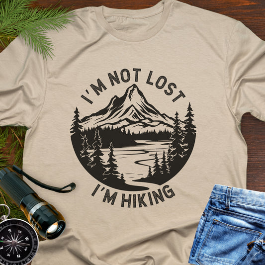 Beige t-shirt with mountain graphic and text 'I'm not lost, I'm hiking' on a wooden surface with outdoor accessories.