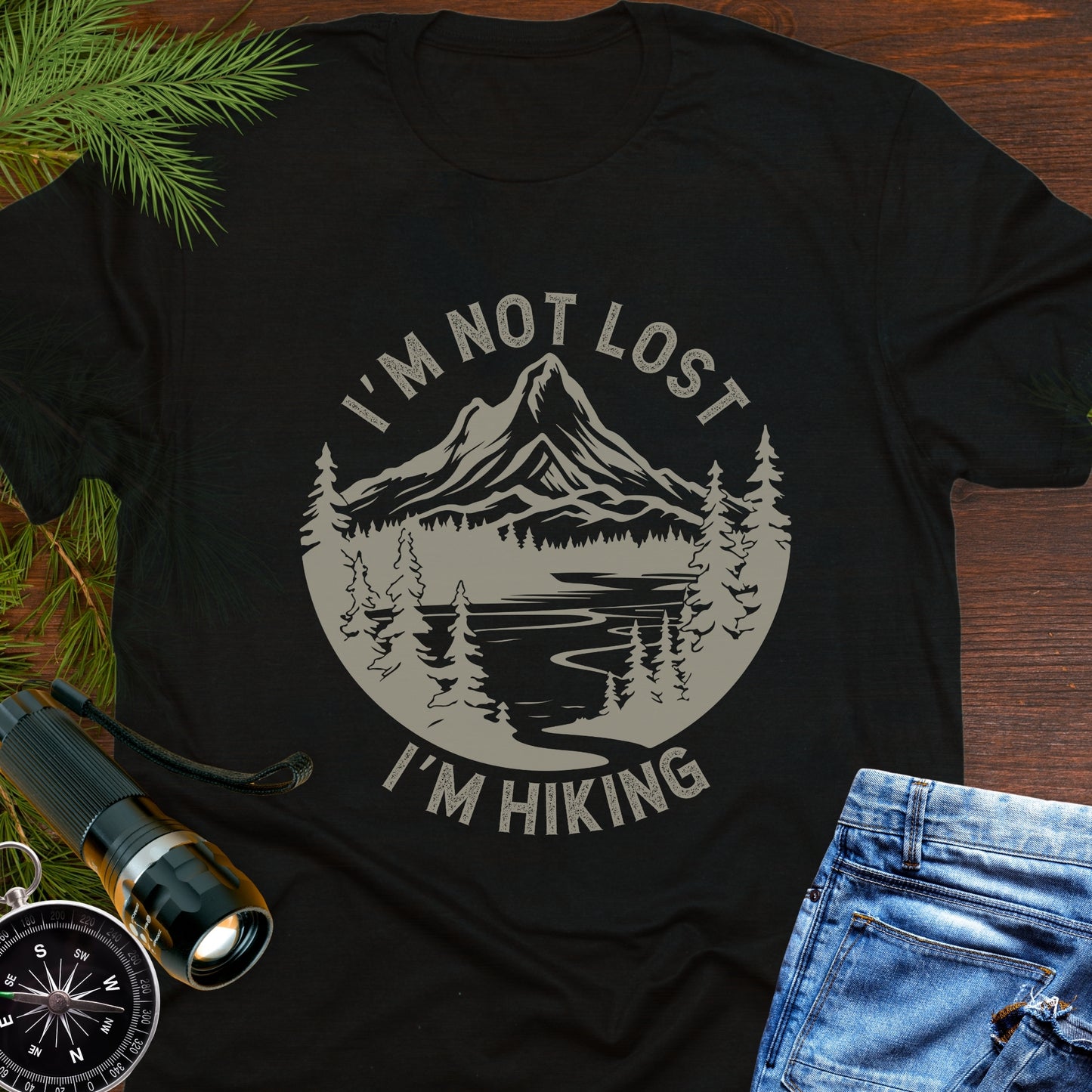 Black t-shirt with 'I'm not lost, I'm hiking' design on a wooden surface with outdoor accessories.