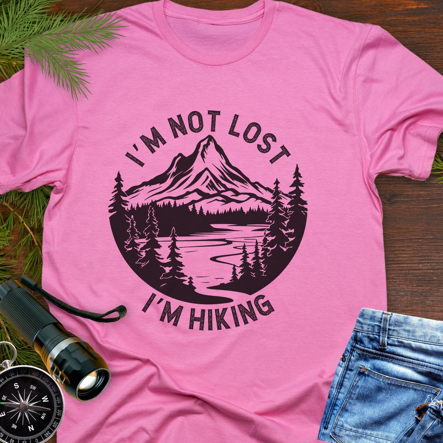 Pink t-shirt with 'I'm not lost, I'm hiking' design on a wooden surface with outdoor accessories.