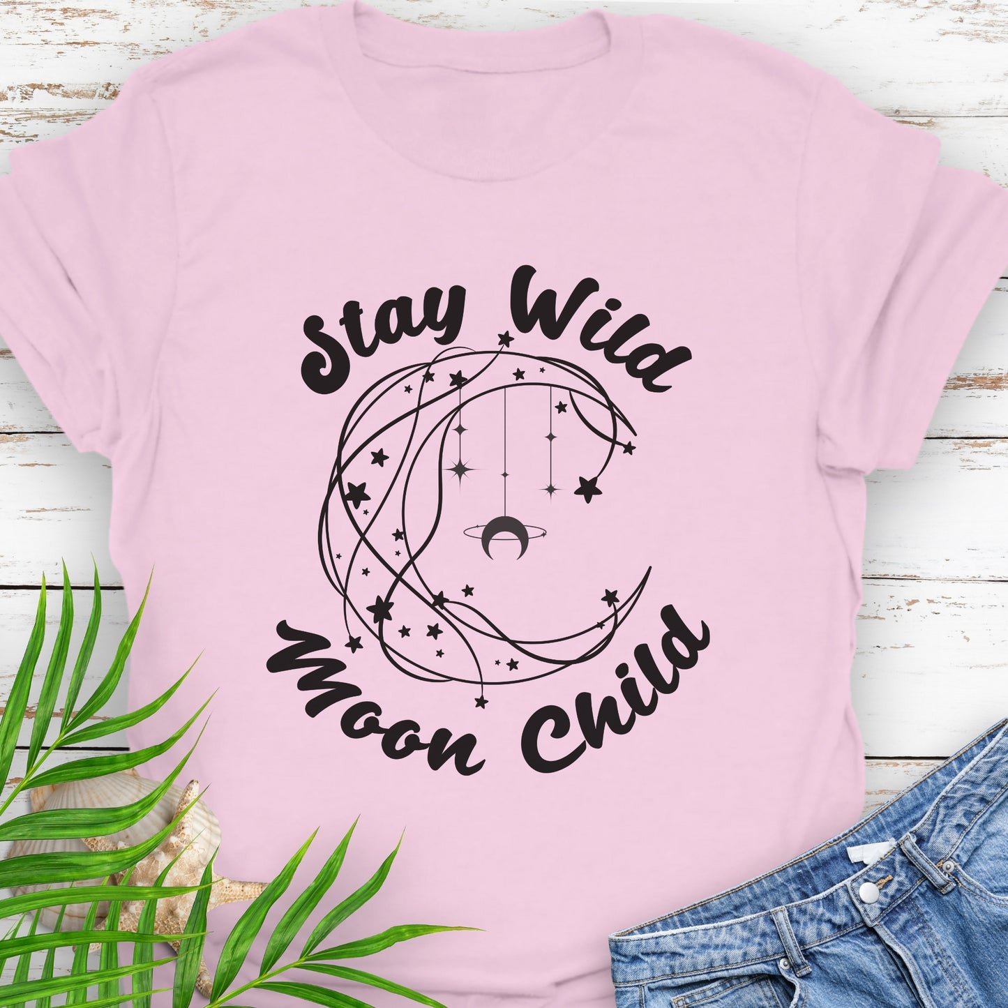 Pink T-shirt with crescent shaped lines and hanging crescent moon and stars that says Stay Wild Moon Child.