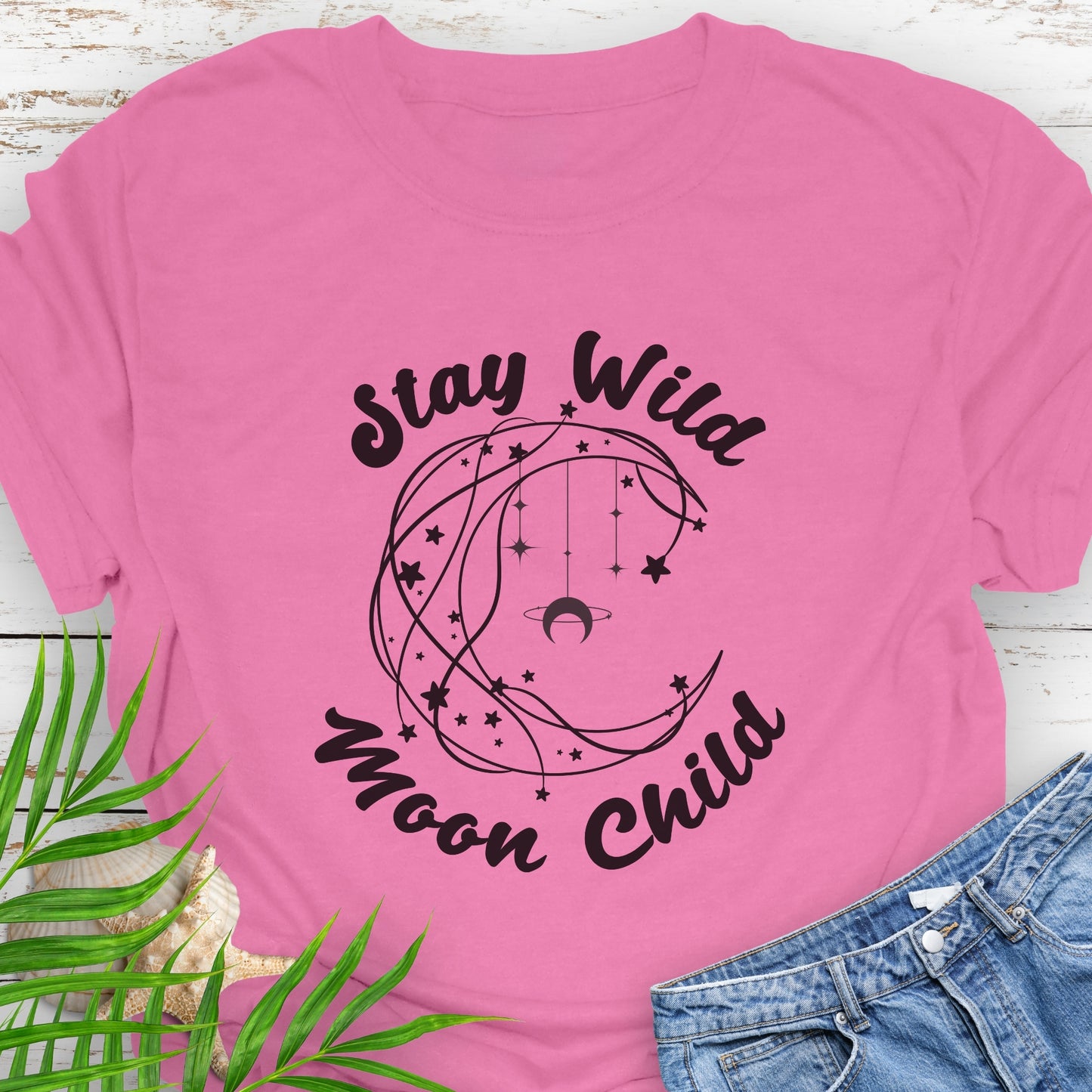 Pink T-shirt with crescent shaped lines and hanging crescent moon and stars that says Stay Wild Moon Child.
