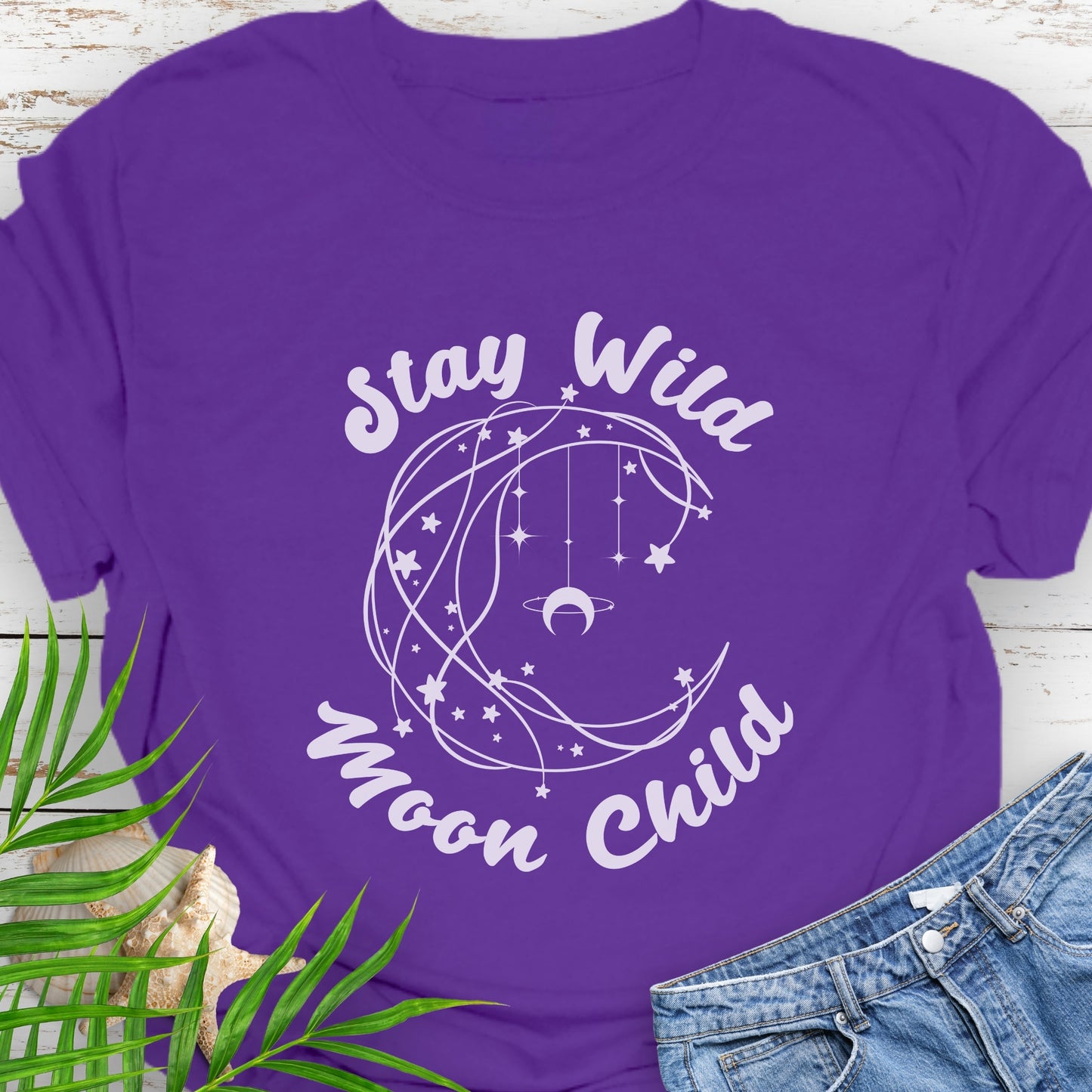 Purple T-shirt with crescent shaped lines and hanging crescent moon and stars that says Stay Wild Moon Child.