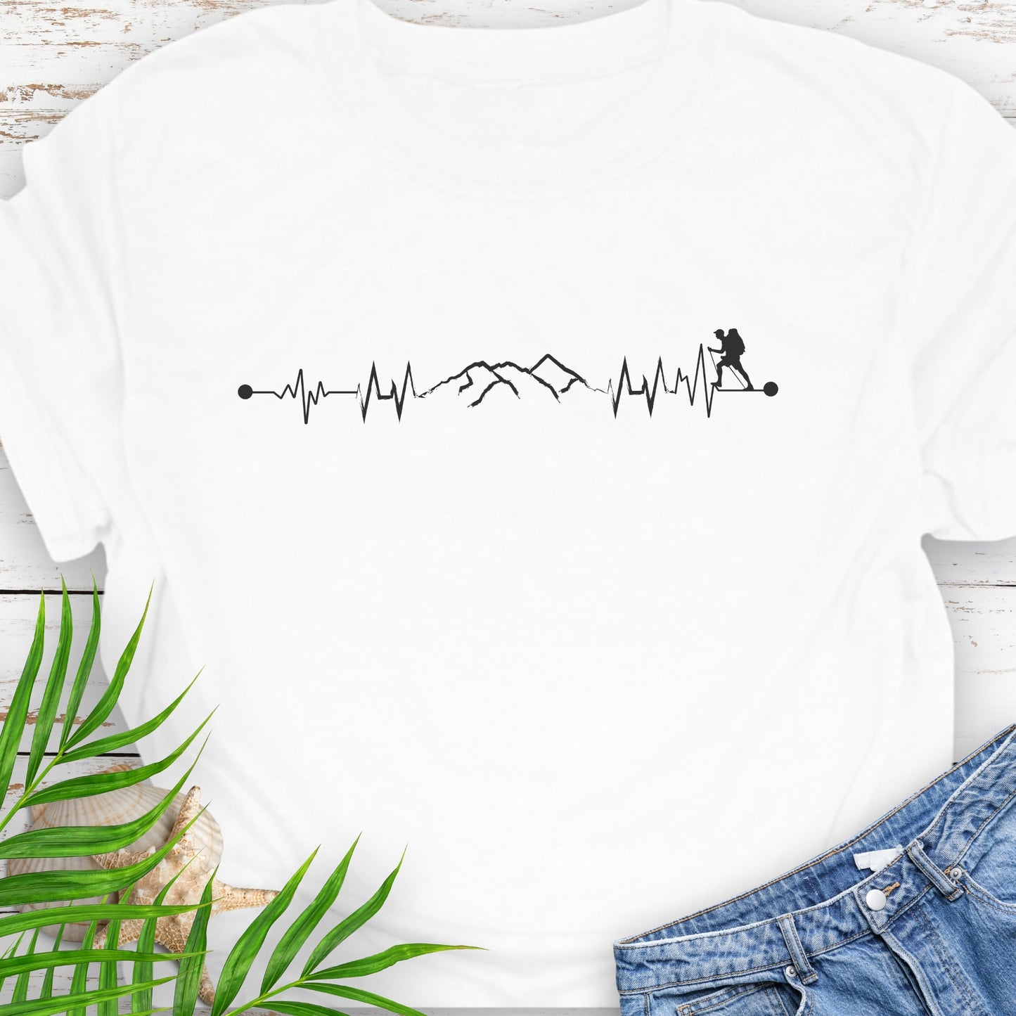 White t-shirt with a black graphic design featuring a heartbeat line, mountains, and a person hiking, placed on a wooden surface with green leaves.