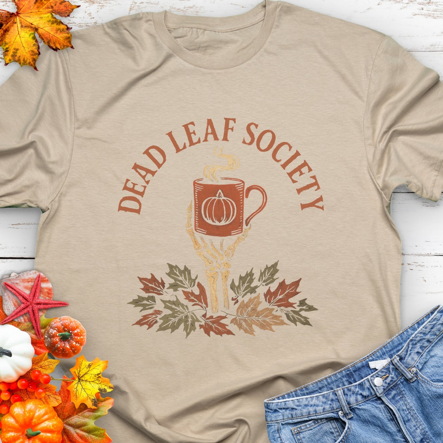 Beige t-shirt with a skeleton hand holding a coffee cup above fall leaves and the text “Dead Leaf Society.”