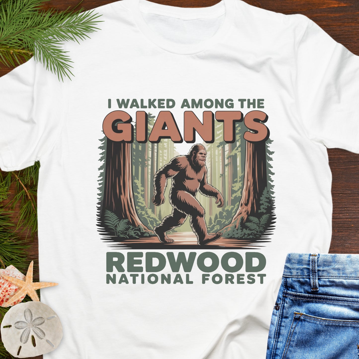 White t-shirt with a Sasquatch graphic and text 'I Walked Among the Giants Redwood National Forest' on a wooden surface.