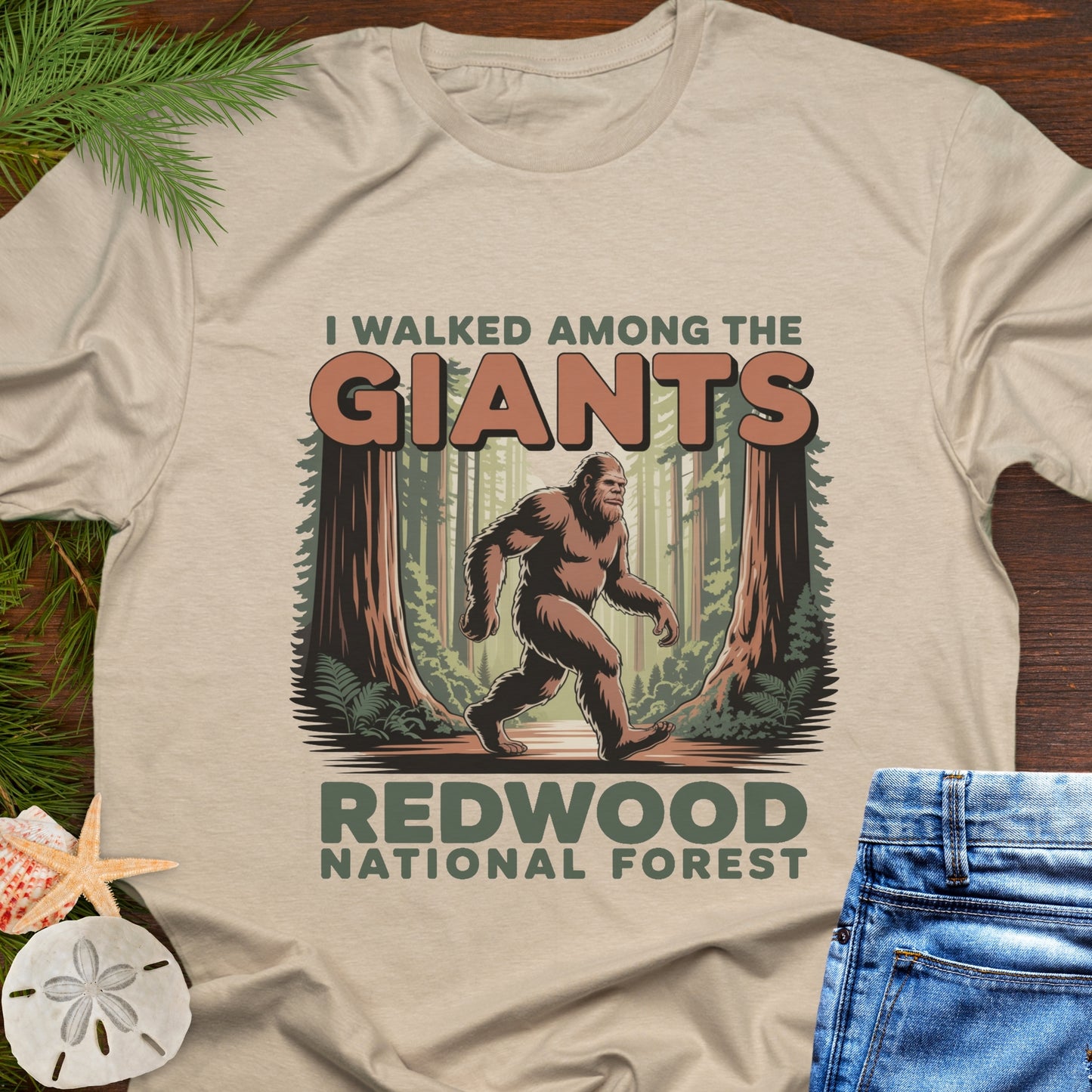 Beige T-shirt with a Bigfoot graphic and text about Redwood National Forest on a wooden surface.