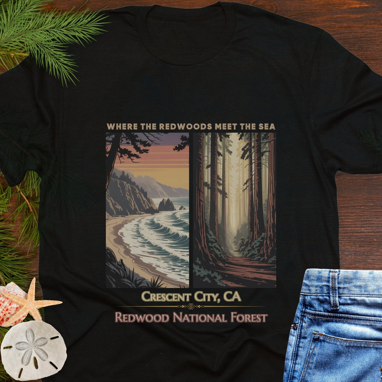 Black t-shirt with a scenic design of Redwood National Forest and beach, placed on a wooden surface with foliage and jeans.