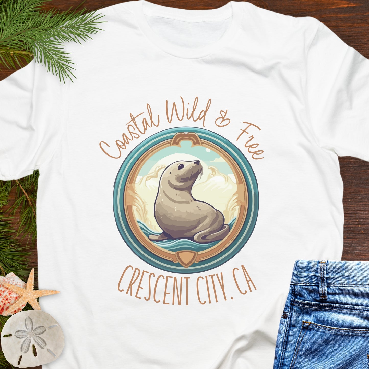 White t-shirt with a seal graphic and text 'Coastal Wild & Free Crescent City, CA' on a wooden surface.