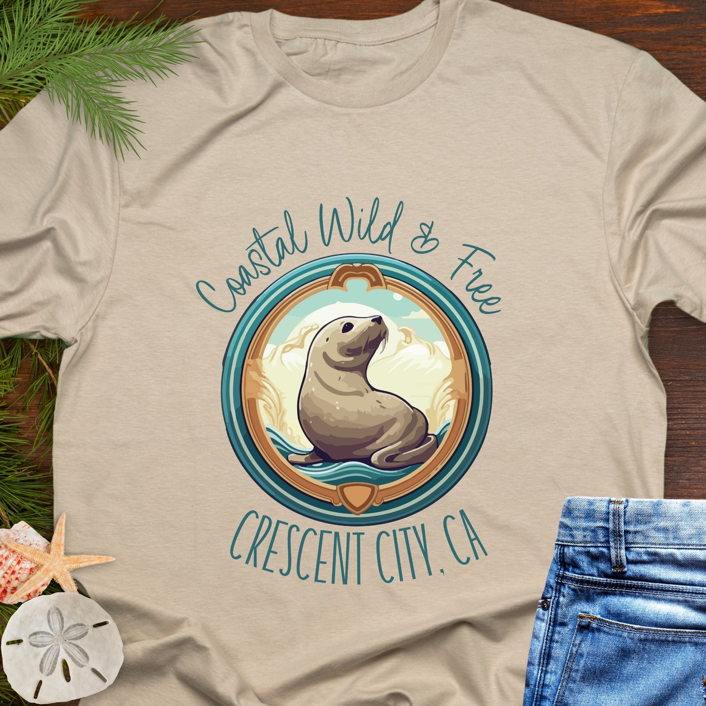 Beige t-shirt with a seal graphic and text 'Coastal Wild & Free Crescent City, CA' on a wooden surface.