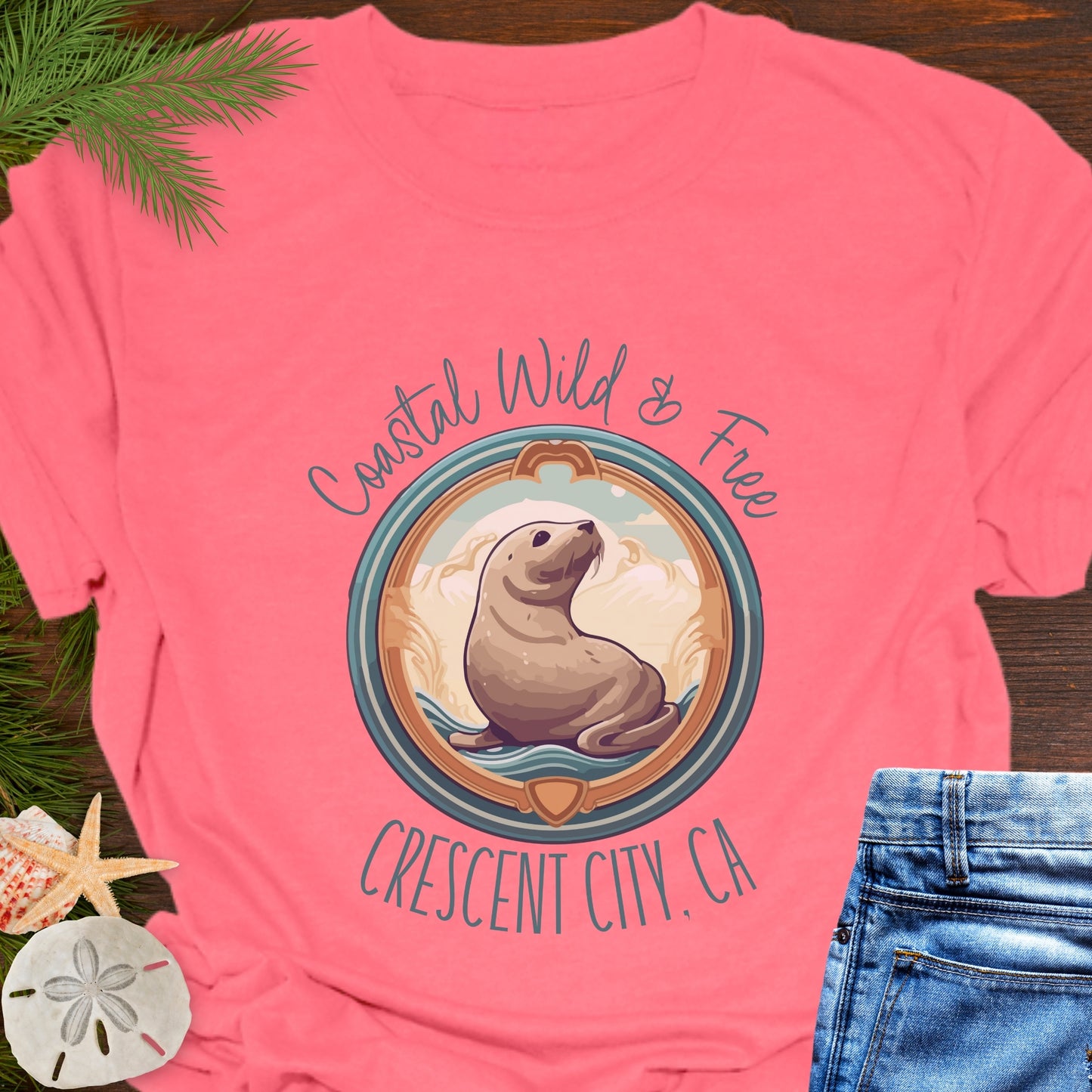 Pink t-shirt with a seal graphic and text on a wooden surface with grass and sand dollars.