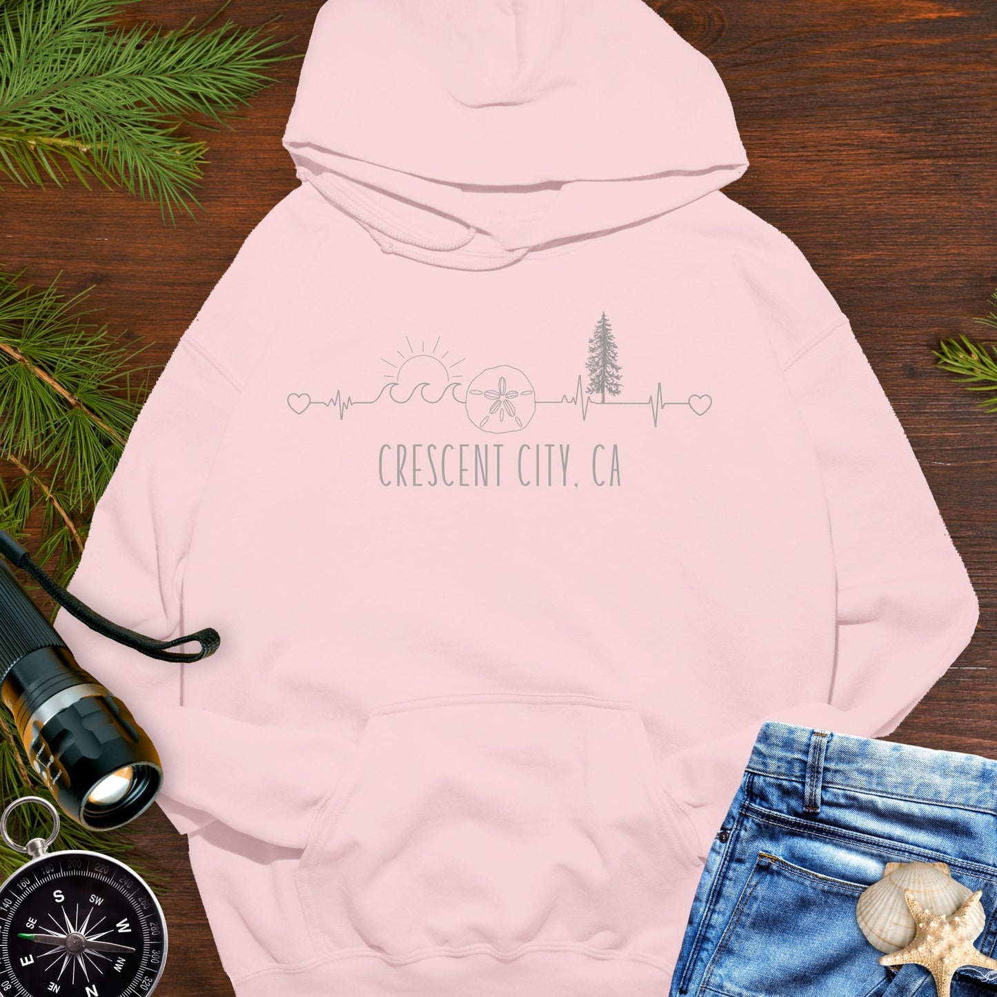 Pink hoodie with a heartline with waves, sand dollar, redwood tree and sun with text 'Crescent City, CA'.