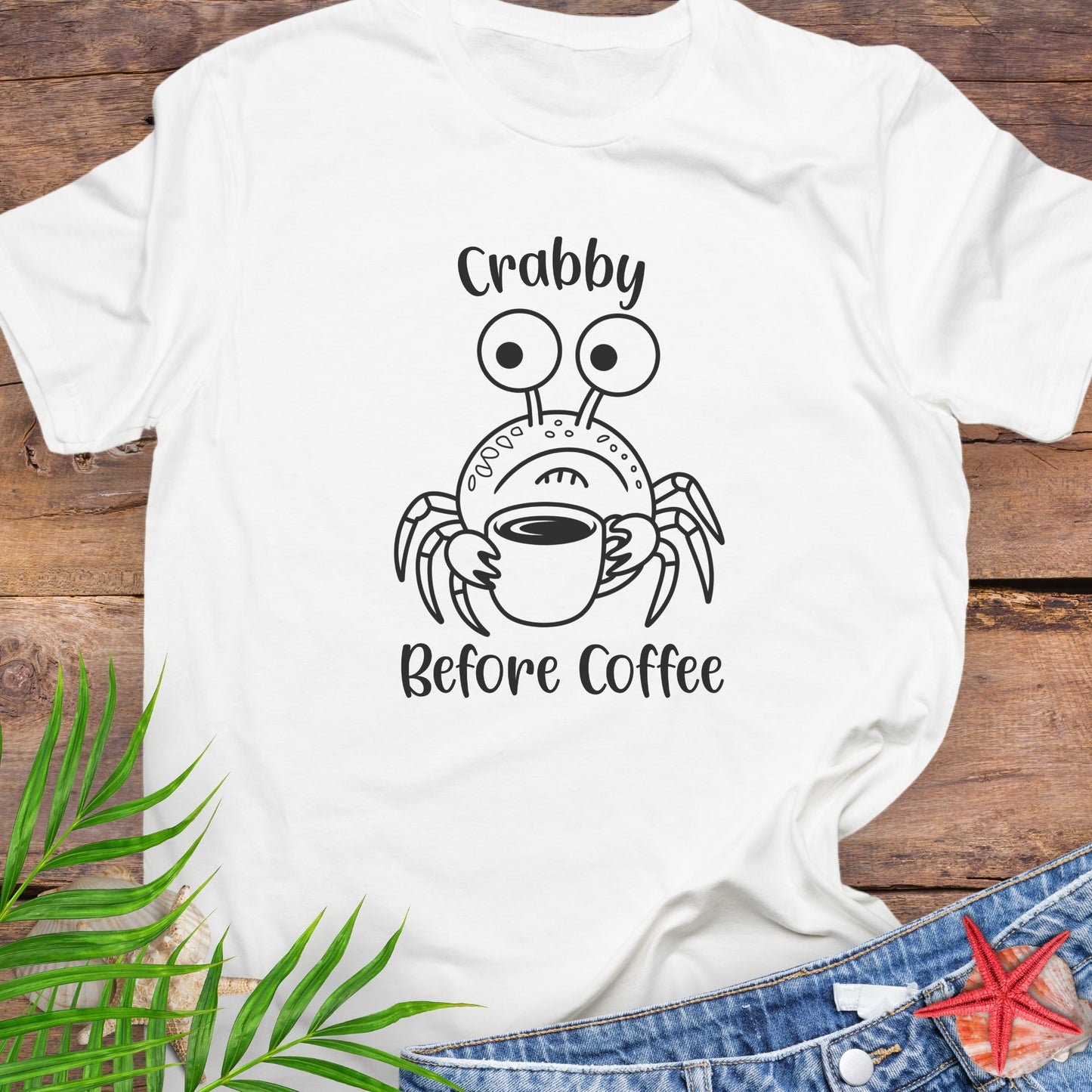 White t-shirt with a crab graphic and 'Crabby Before Coffee' text on a wooden surface with leaves and jeans.