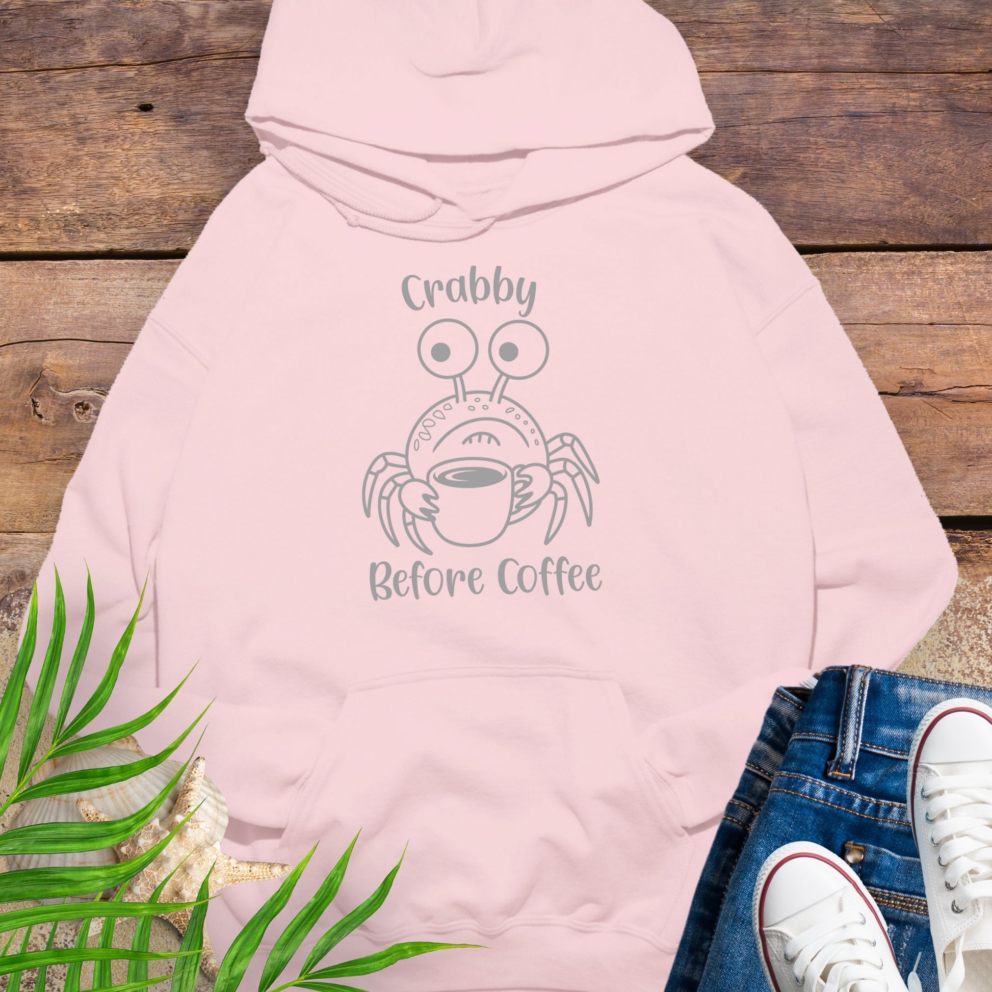 Pink hoodie with crab graphic and text 'Crabby Before Coffee' on a wooden surface with jeans and sneakers.