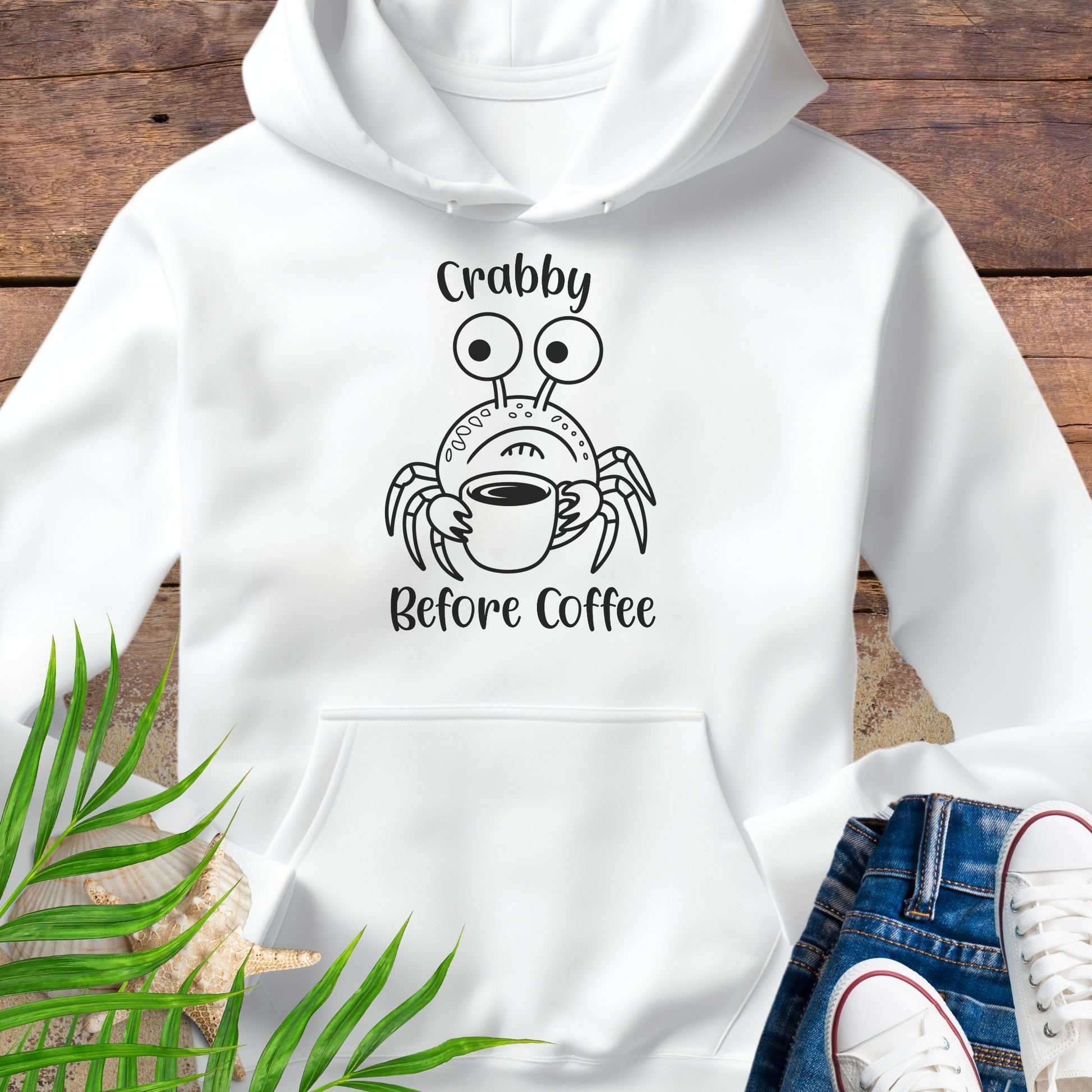 White hoodie with a crab graphic and text 'Crabby Before Coffee' on a wooden surface.