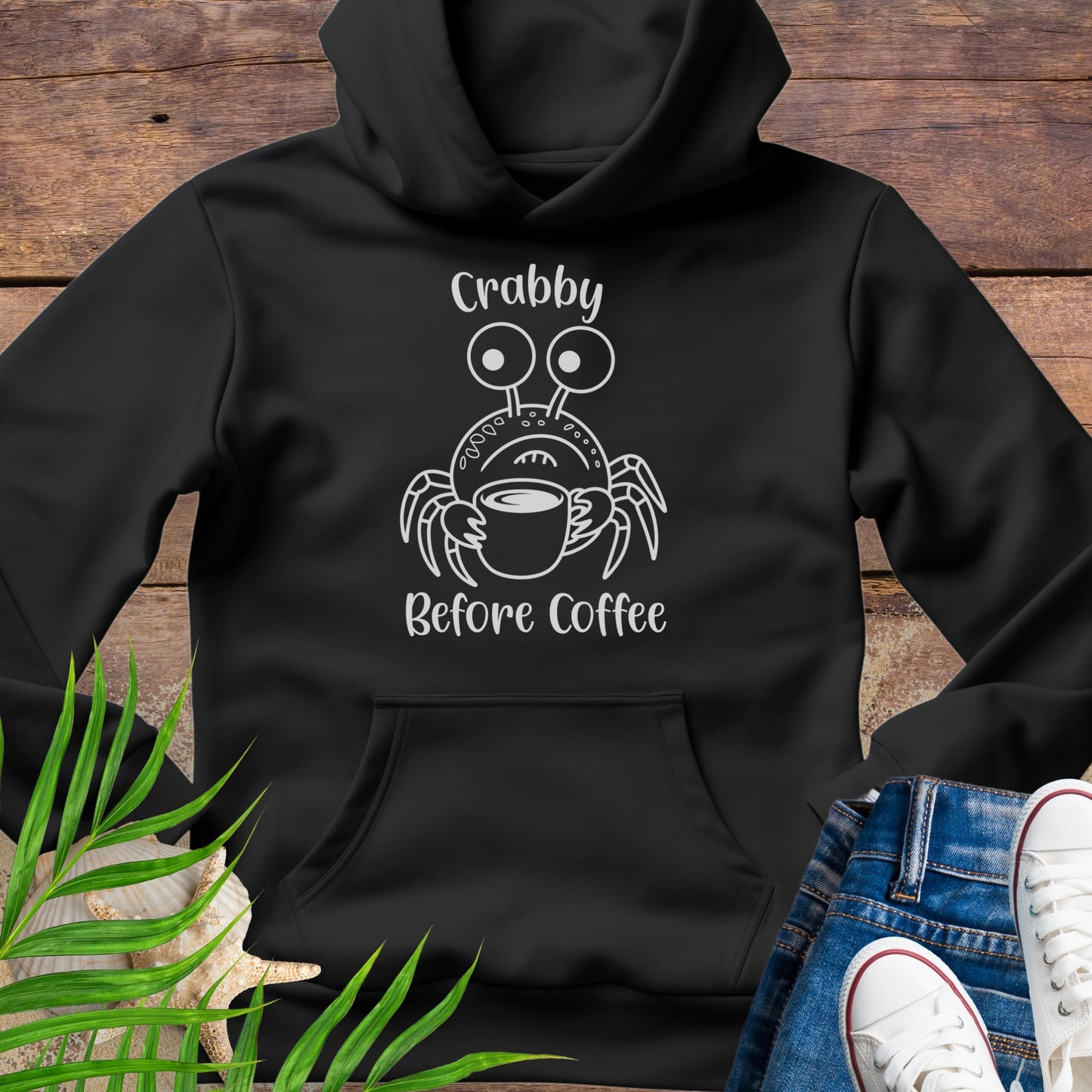 Black hoodie with a crab graphic and text 'Crabby Before Coffee' on a wooden surface.