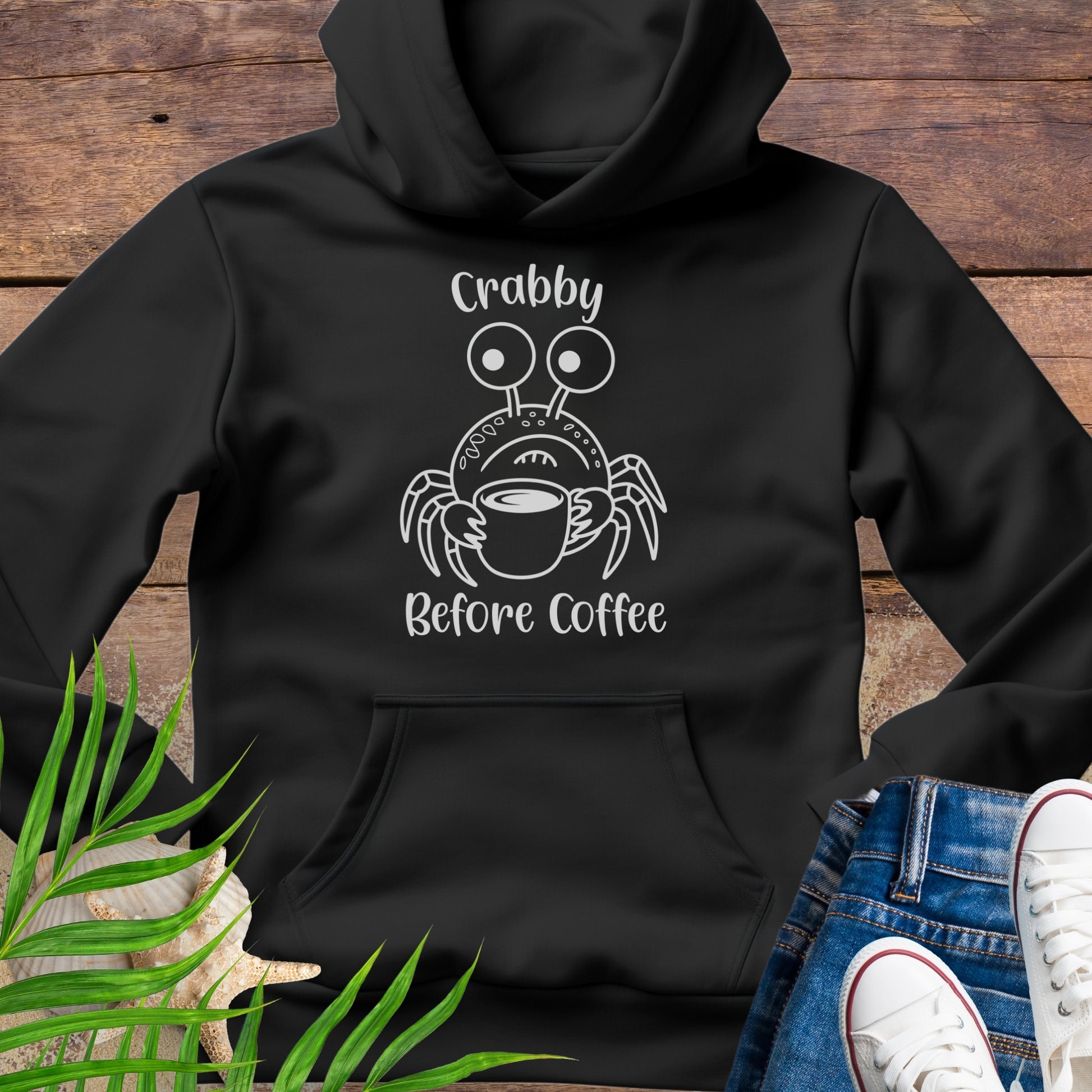 Black hoodie with a crab graphic and text 'Crabby Before Coffee' on a wooden surface.