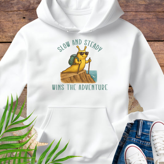 White hoodie with a snail graphic and text 'Slow and Steady Wins the Adventure' on a wooden surface.
