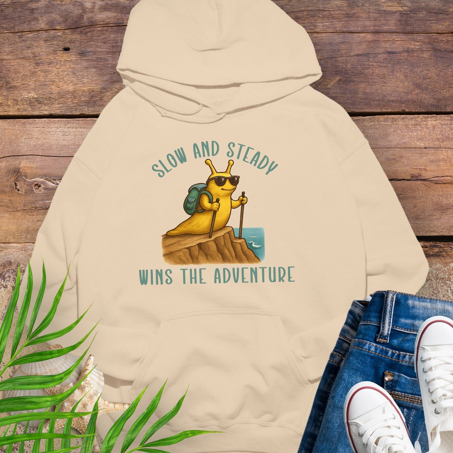 Beige hoodie with a snail graphic and text 'Slow and Steady Wins the Adventure' on a wooden background.